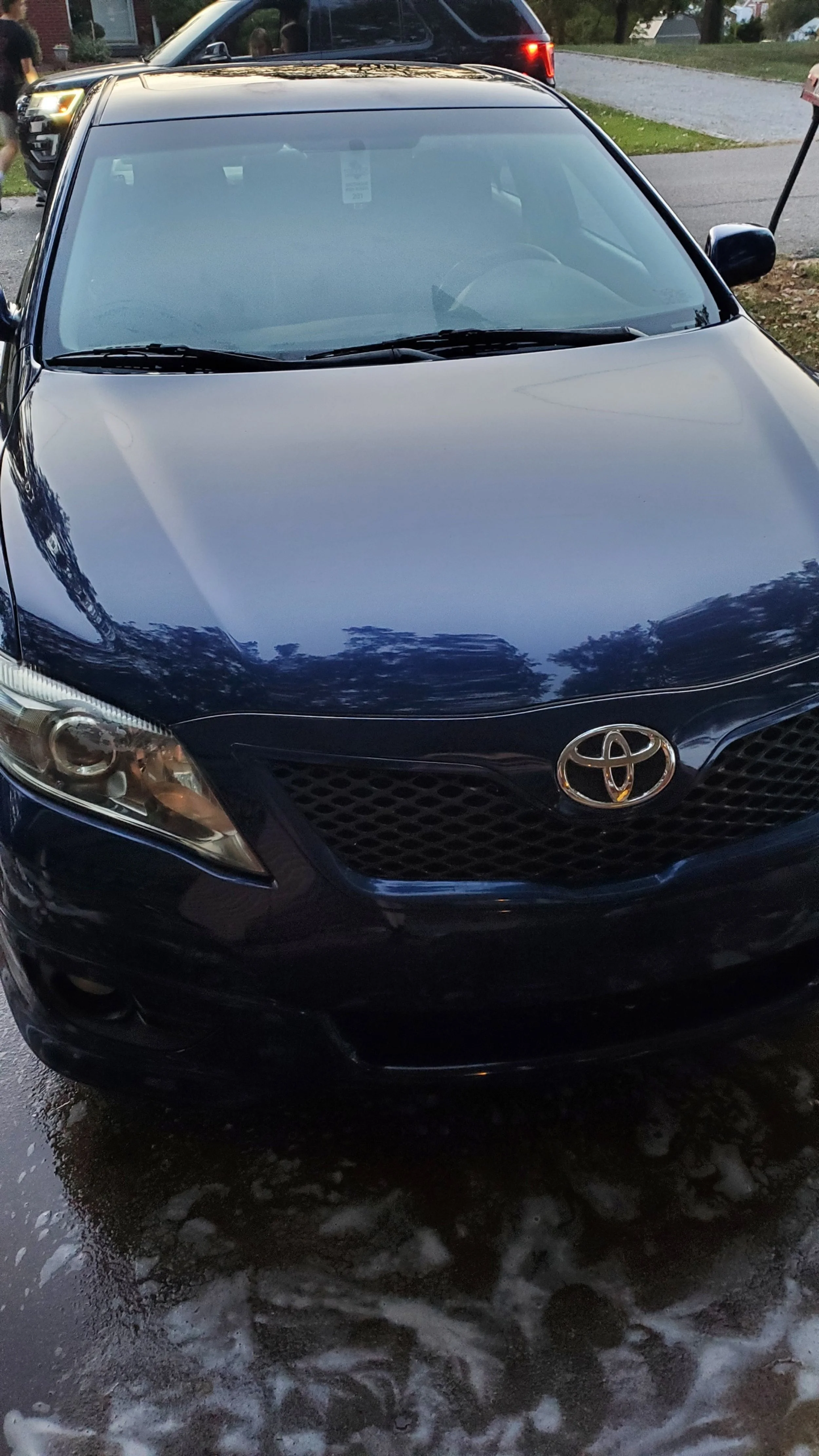 Black Toyota sedan parked outdoors on wet ground with soap suds, greenery, and other vehicles in the background.