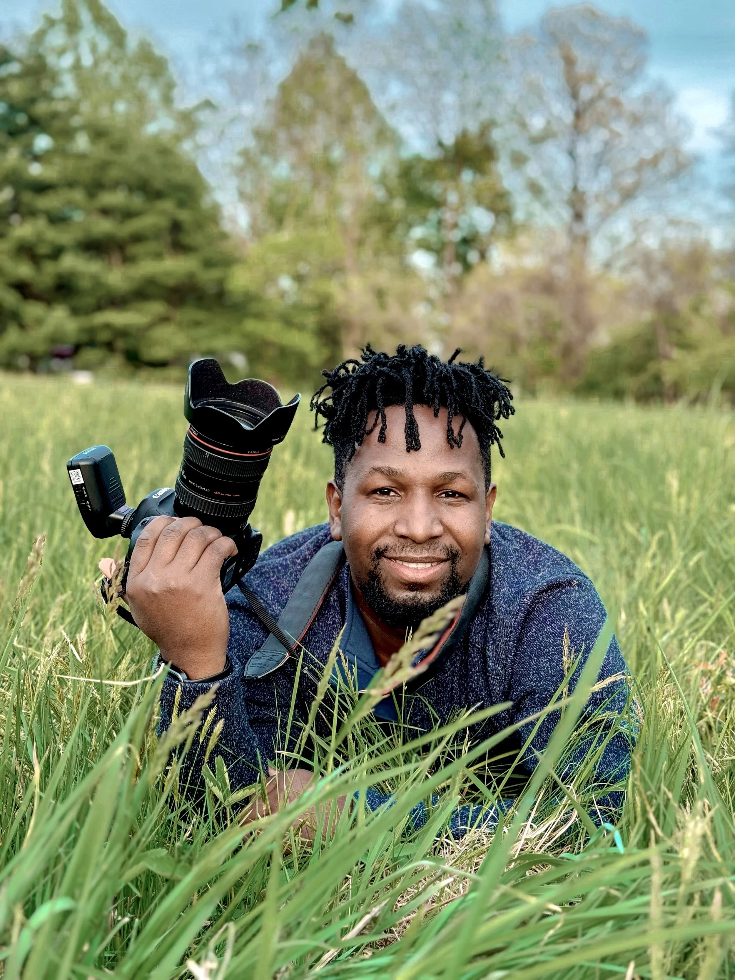A man with black twisted hair and a beard lying in a grassy field during daytime, holding a professional camera with a large lens, smiling at the camera.