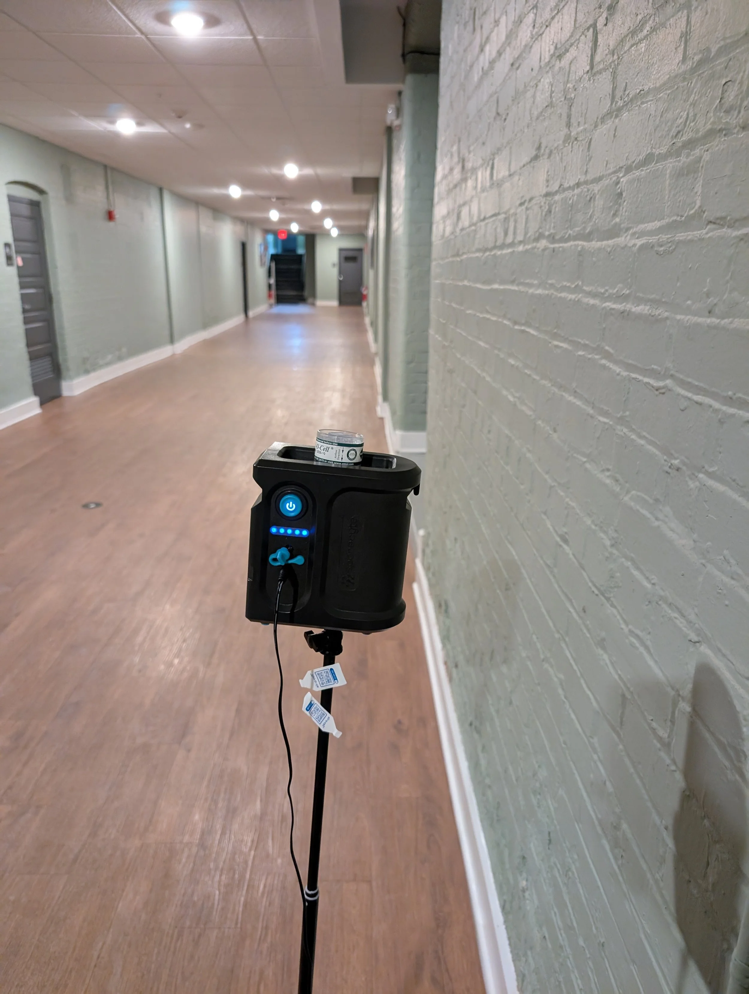 Empty hallway with brick walls and wood flooring, lit by ceiling lights, with a black medical or medical-related device on a stand in the foreground.