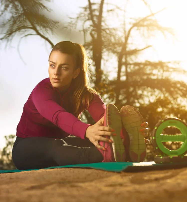 Young woman in athletic clothing stretching outdoors on a yoga mat, with trees and sunlight in the background during sunset.