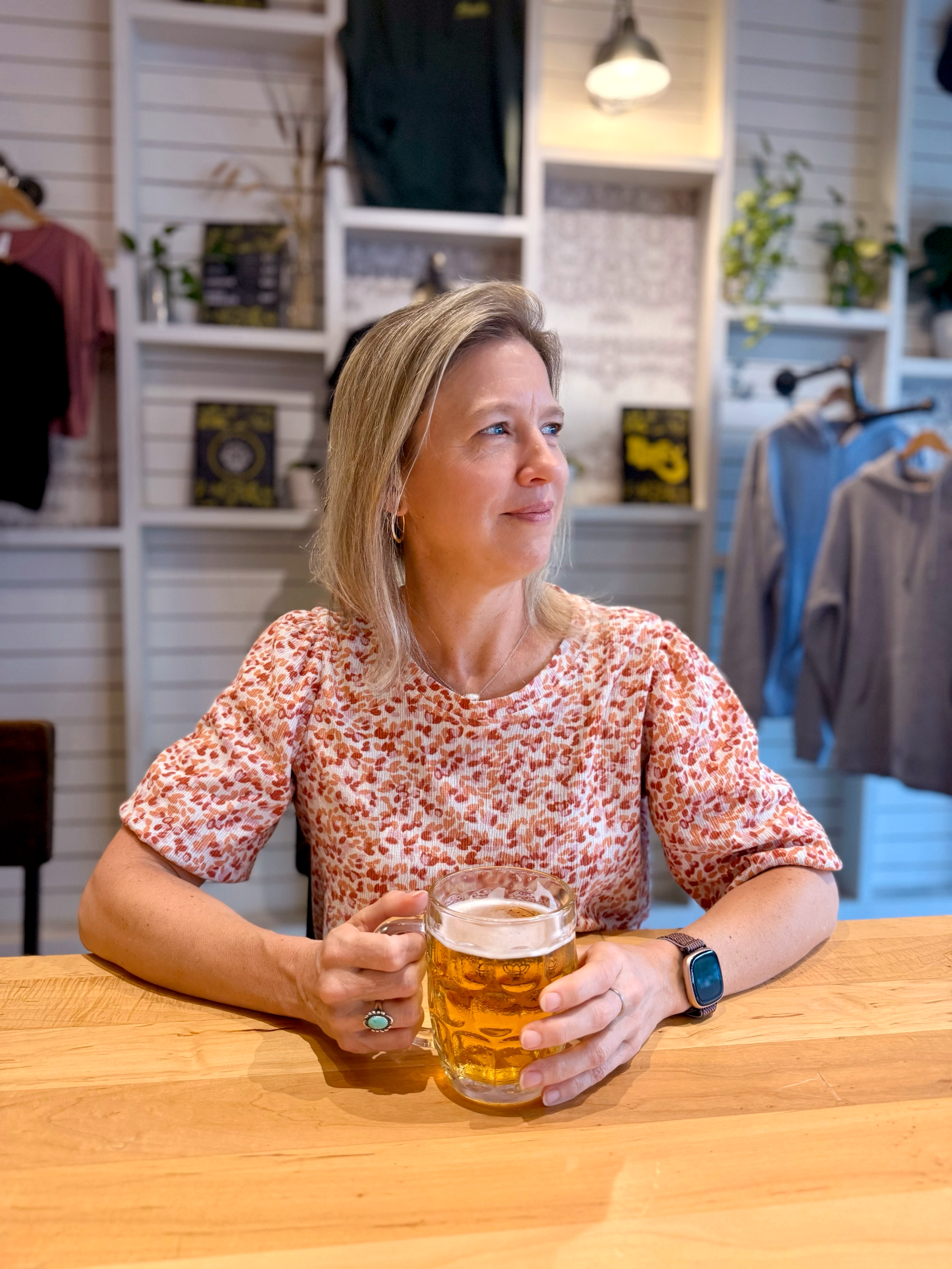 Julie Mastbrook, a woman and designer, sitting at a wooden table with a mug of beer in hand looking out the window.