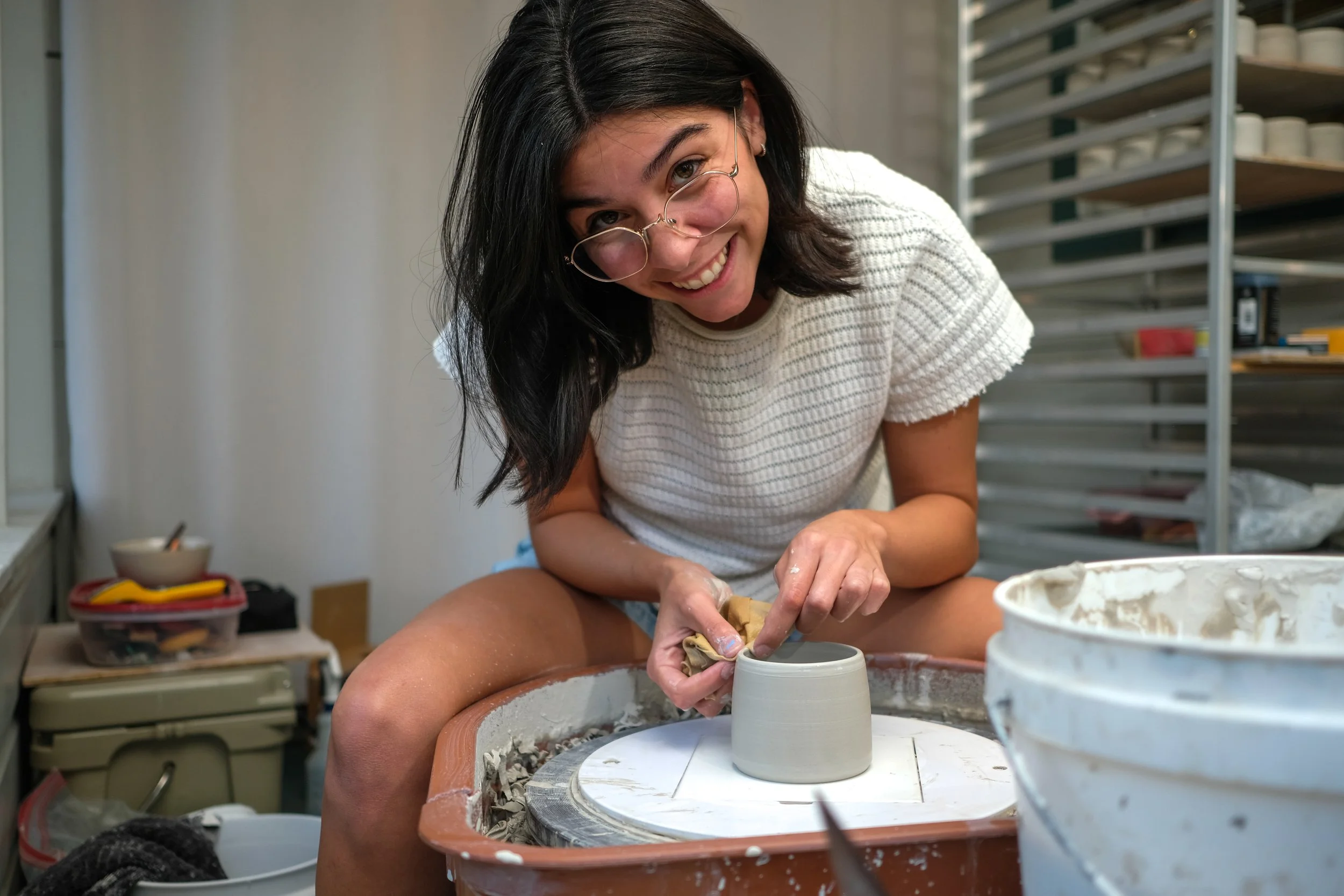Young woman smiling while shaping a ceramic mug on a pottery wheel in a studio.