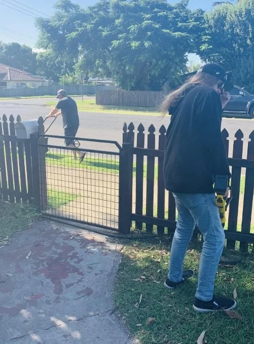 A woman standing near a black picket fence talking on her phone, with a man gardening with a rake on the other side of the fence on a sunny day.