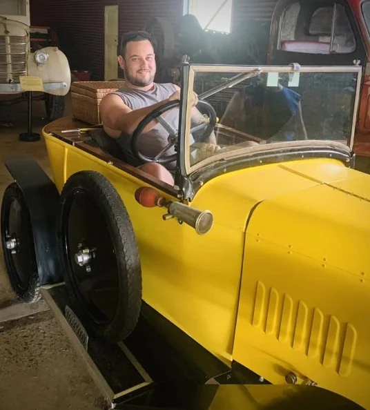 A man sitting inside a small, yellow, vintage-style car with a clear windshield, parked indoors.