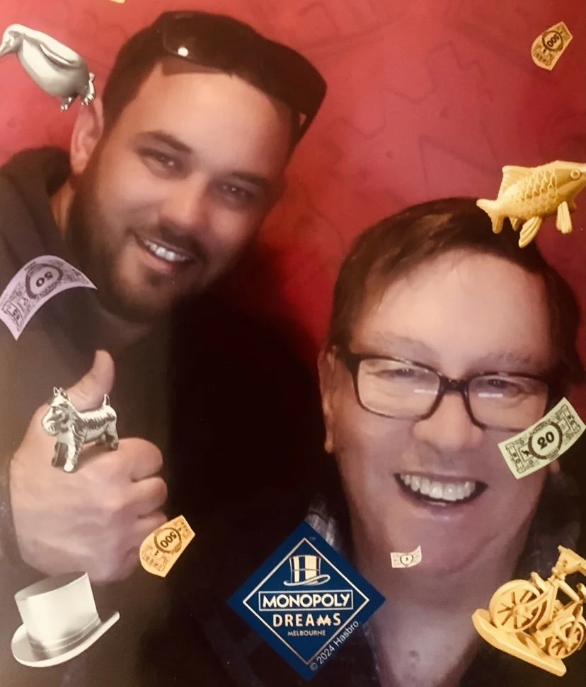 Two men smiling and taking a selfie with Monopoly-themed decoration and stickers, including Monopoly money and game pieces, against a red background.