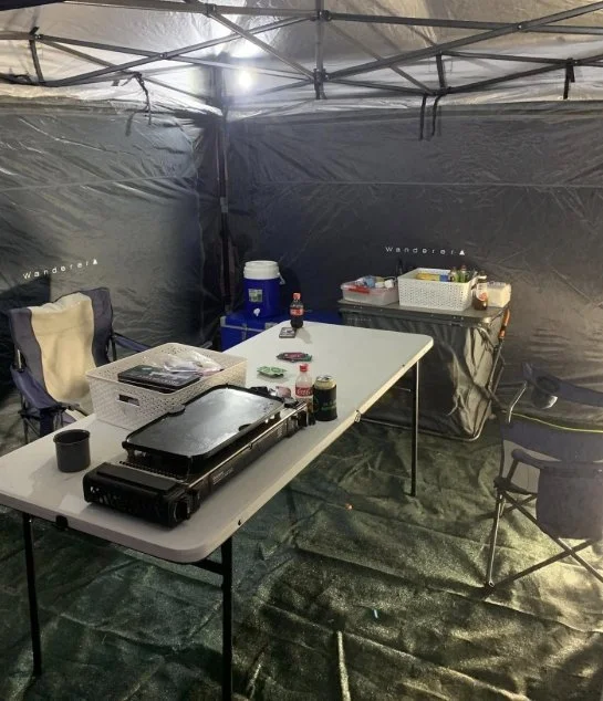 Camping setup inside a large tent with folding table holding a portable grill, drinks, and supplies; chairs and a cooler also visible.