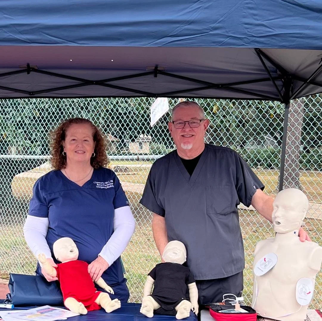 Two emergency medical professionals, a woman and a man, standing under a canopy tent outdoors with medical training mannequins on a table in front of them.