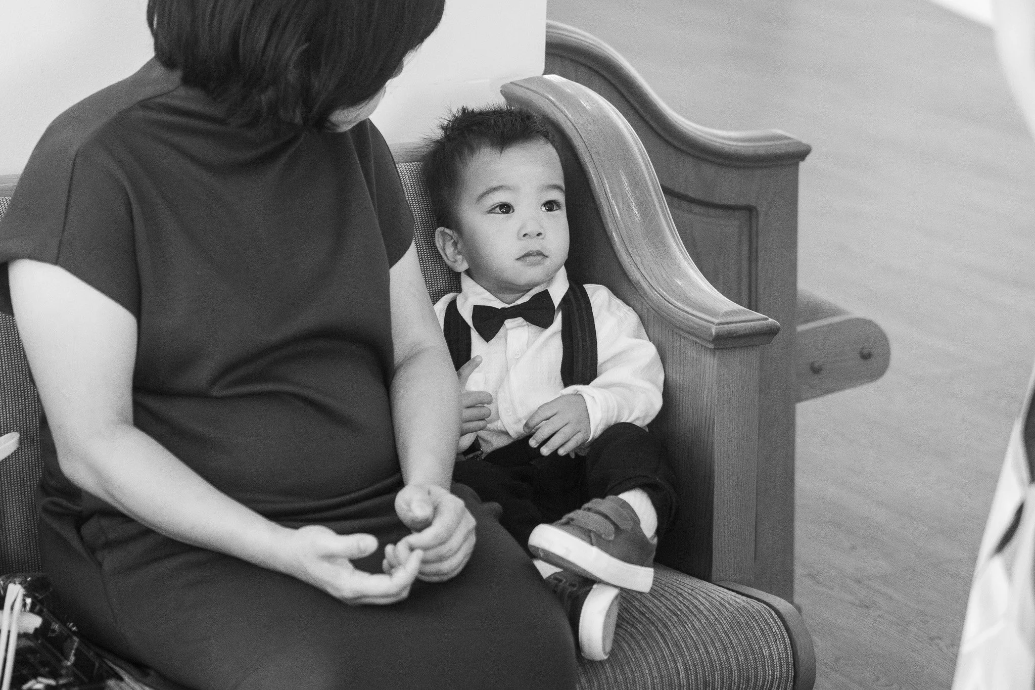 A young boy dressed in a formal shirt, vest, and bow tie sitting on a church pew, looking slightly upwards, while a woman sitting next to him is engaging with him, visible only from the neck down in the image.