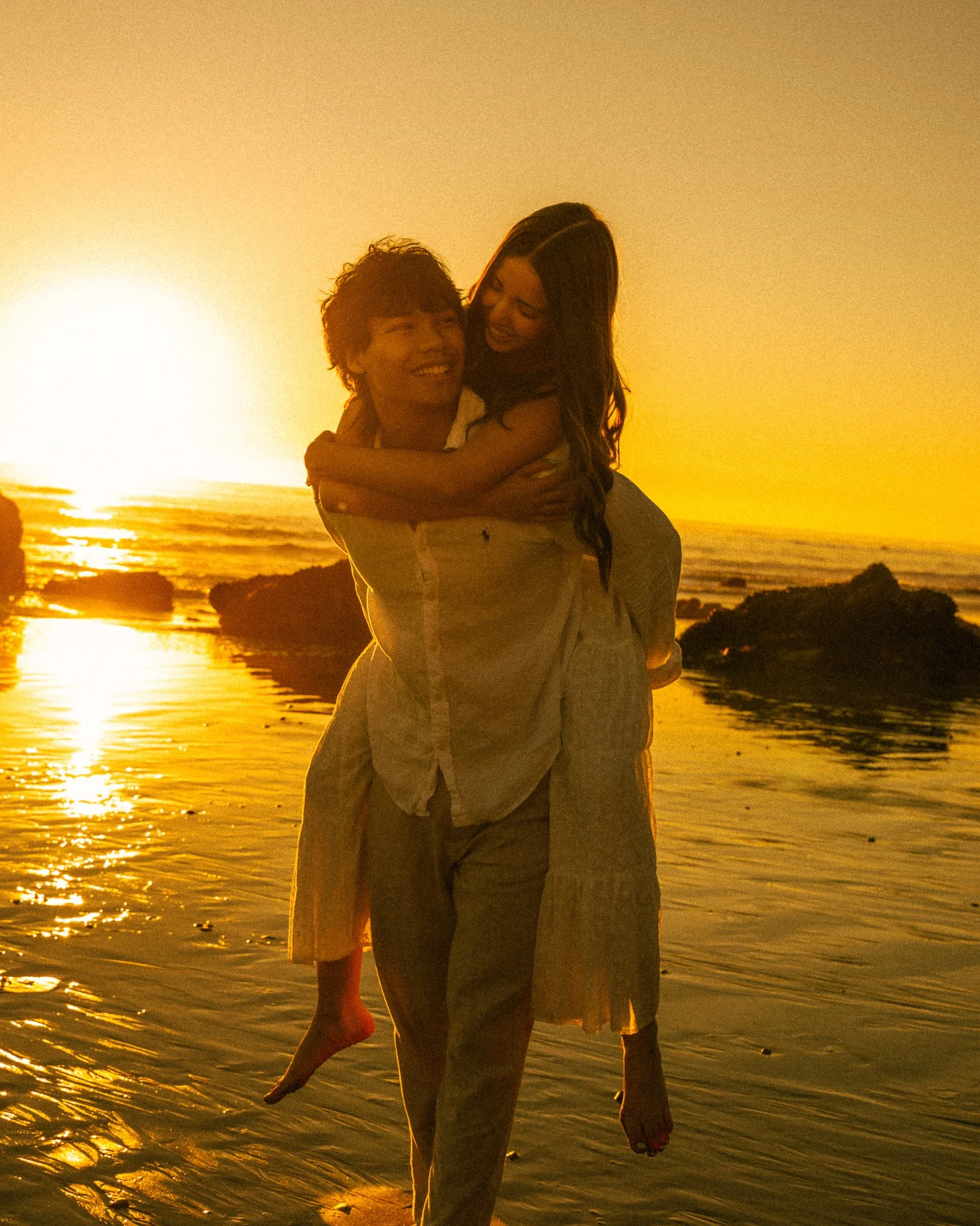 A couple smiling and embracing on the beach at sunset, with the ocean and rocks in the background.