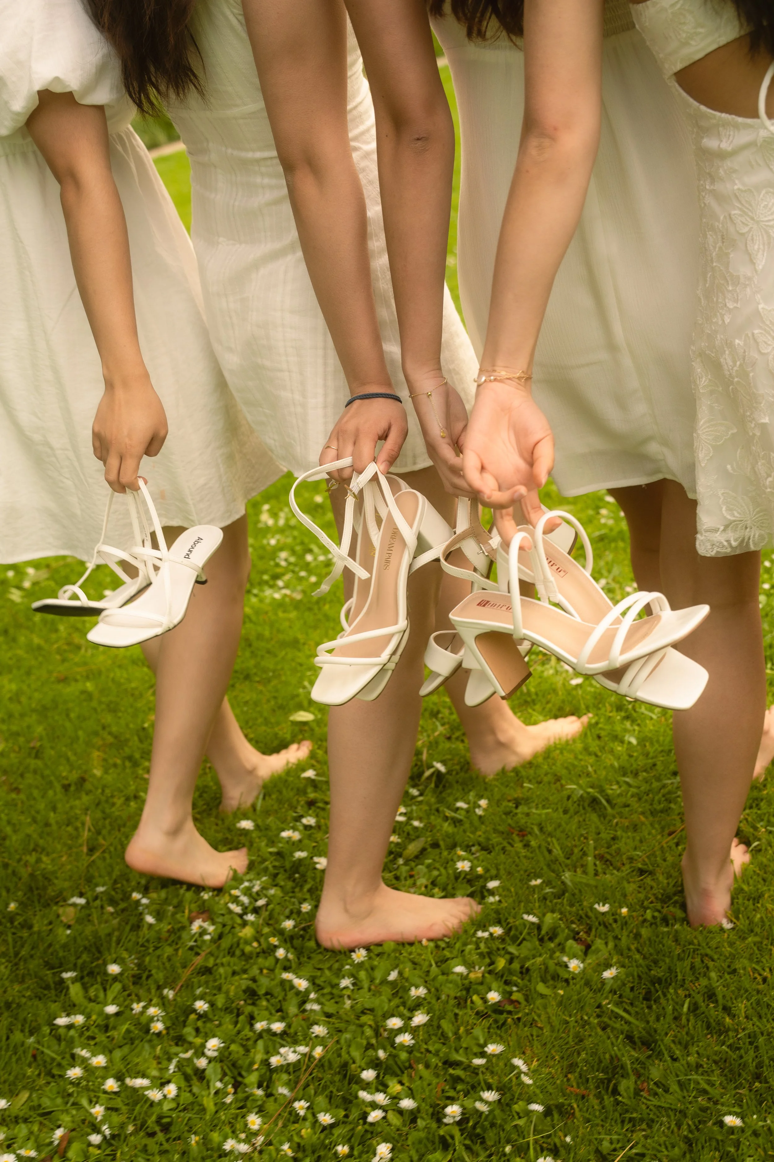 Four women standing barefoot on grass, holding white high-heeled shoes, all dressed in white dresses, with small white flowers on the grass.