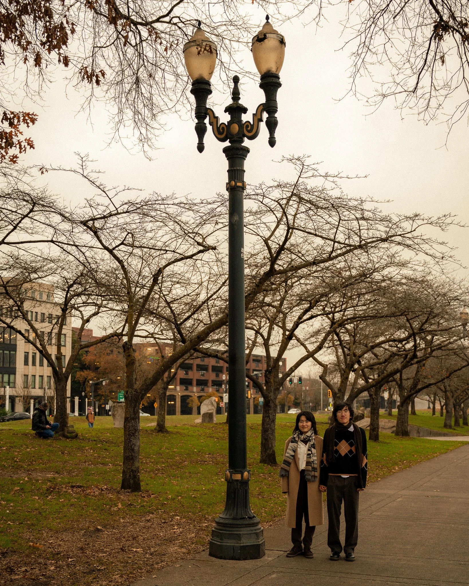 Two women standing close together beneath a decorative black street lamp in a park with leafless trees and buildings in the background. One is wearing a light-colored coat and scarf, and the other a dark sweater and glasses.