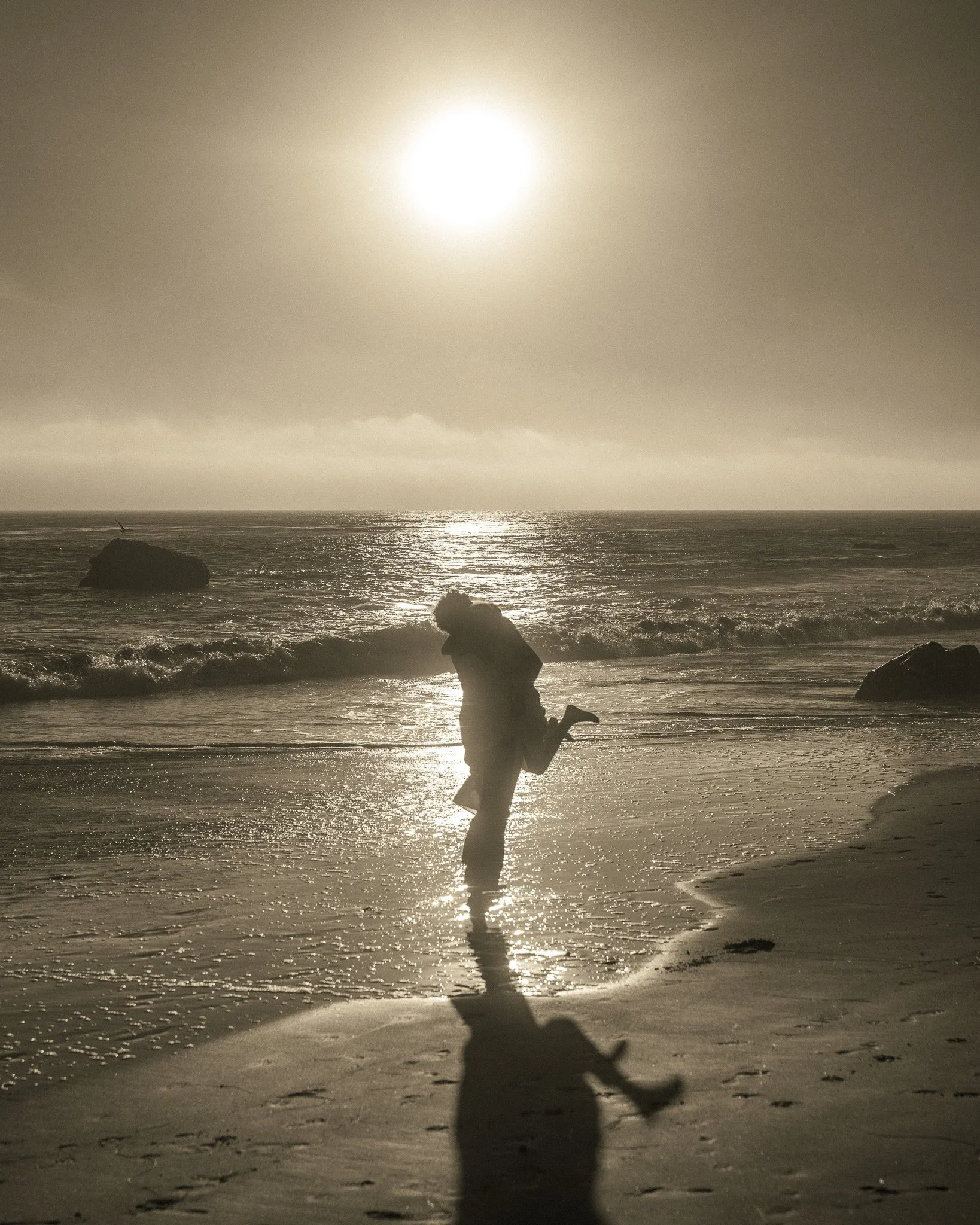 A person kneeling on the beach during sunset, with the sun shining brightly above the horizon and reflecting on the water.
