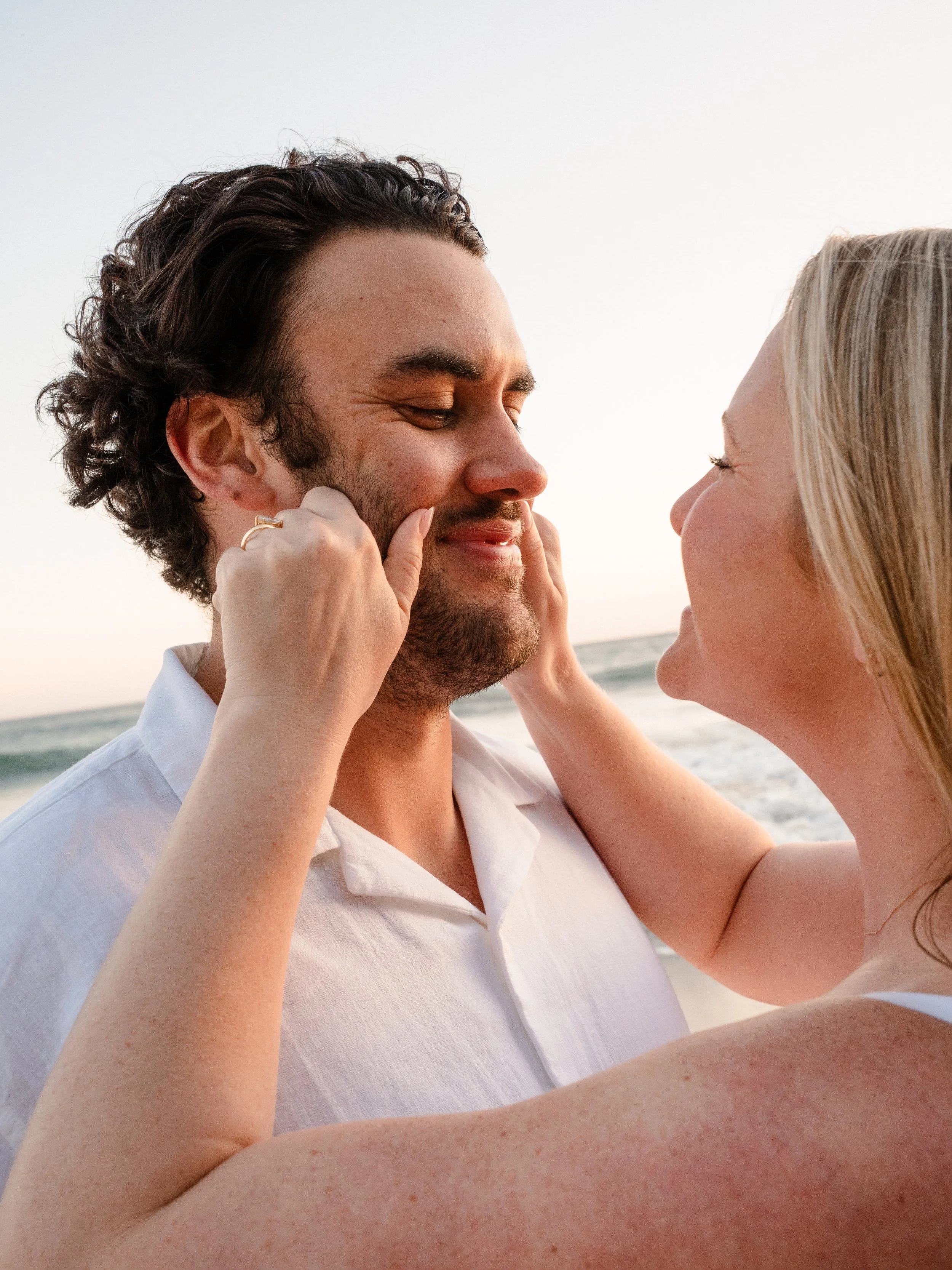 A smiling man with dark curly hair and a beard being gently touched on his face by a blonde woman at the beach during sunset.