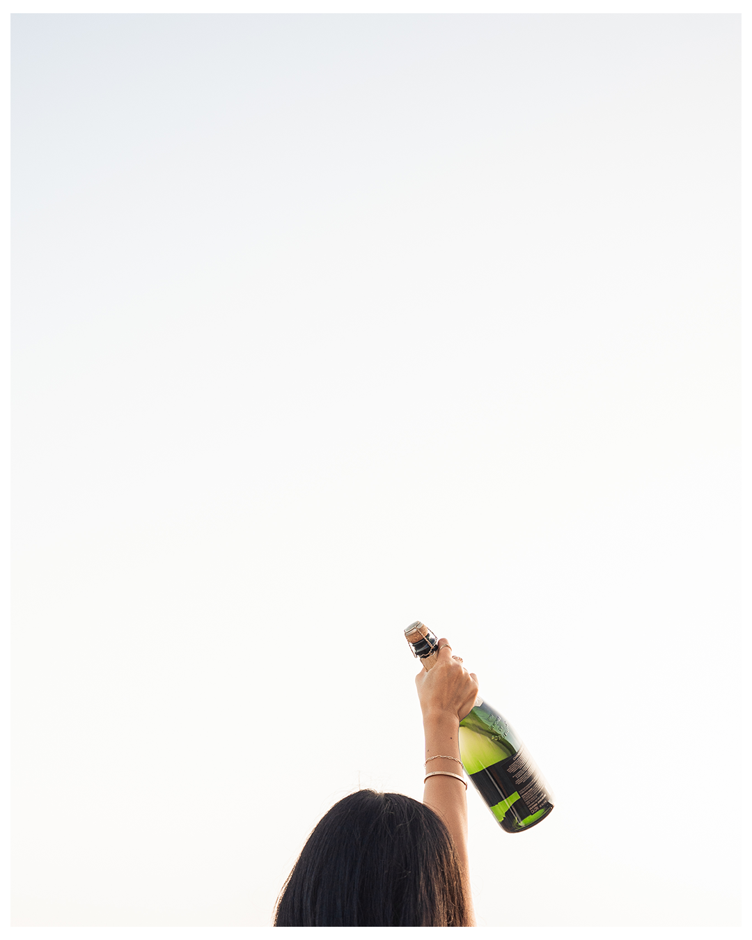Person with long dark hair holding a champagne bottle in the air against a clear sky.
