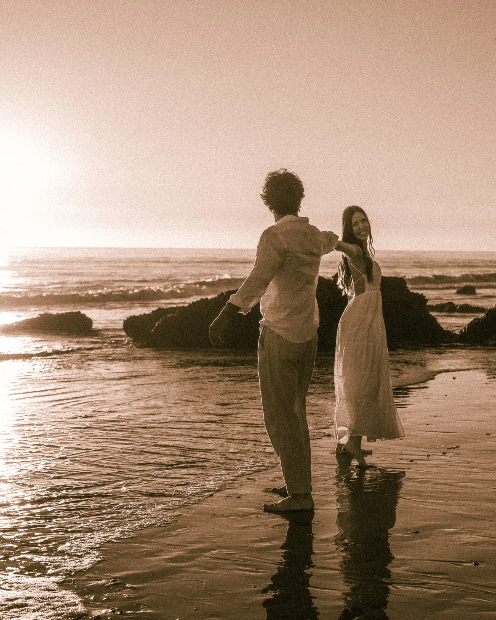 A man and woman stand on a beach at sunset, holding hands and smiling at each other, with ocean waves and rocks in the background.