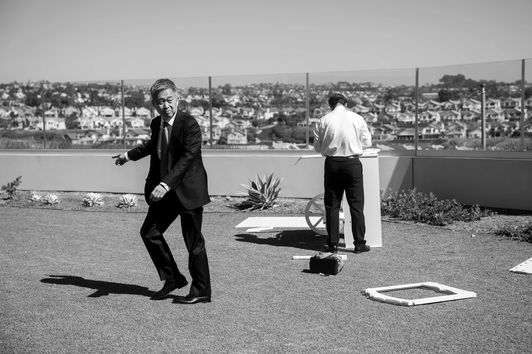 Two men in business attire working on a rooftop, with one walking and the other facing away, amid construction or setup materials, overlooking a residential area.