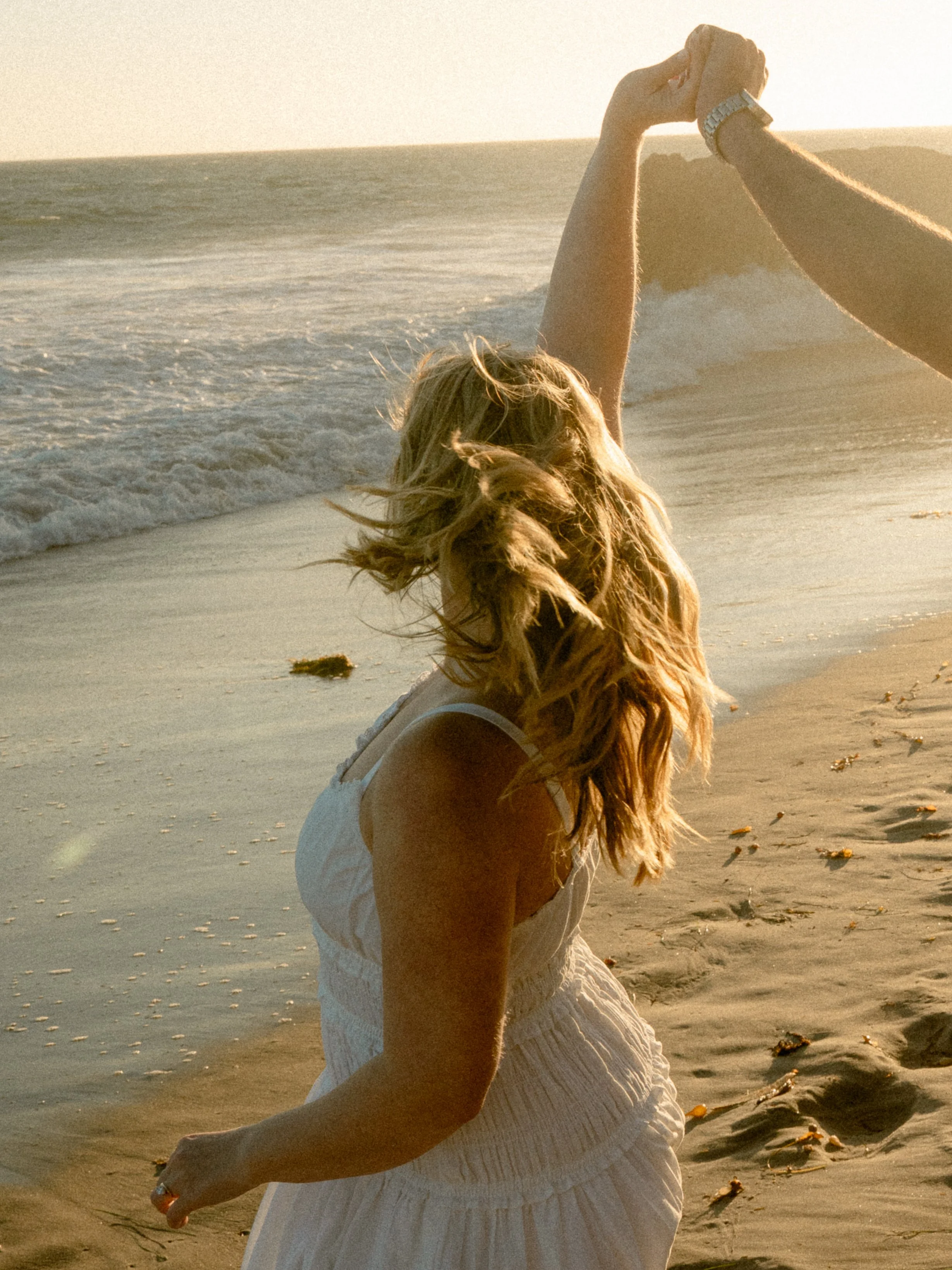 A woman with long blonde hair in a white dress sitting on a sandy beach, holding hands with a person across from her, during sunset.
