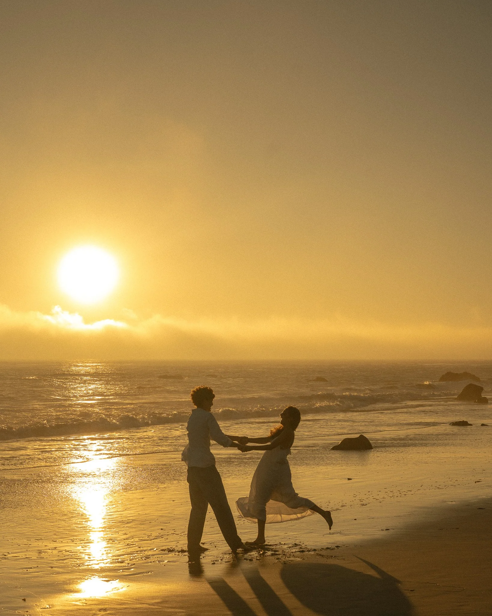 A couple holding hands and playing on the beach at sunset, with the ocean and a cloudy sky in the background.