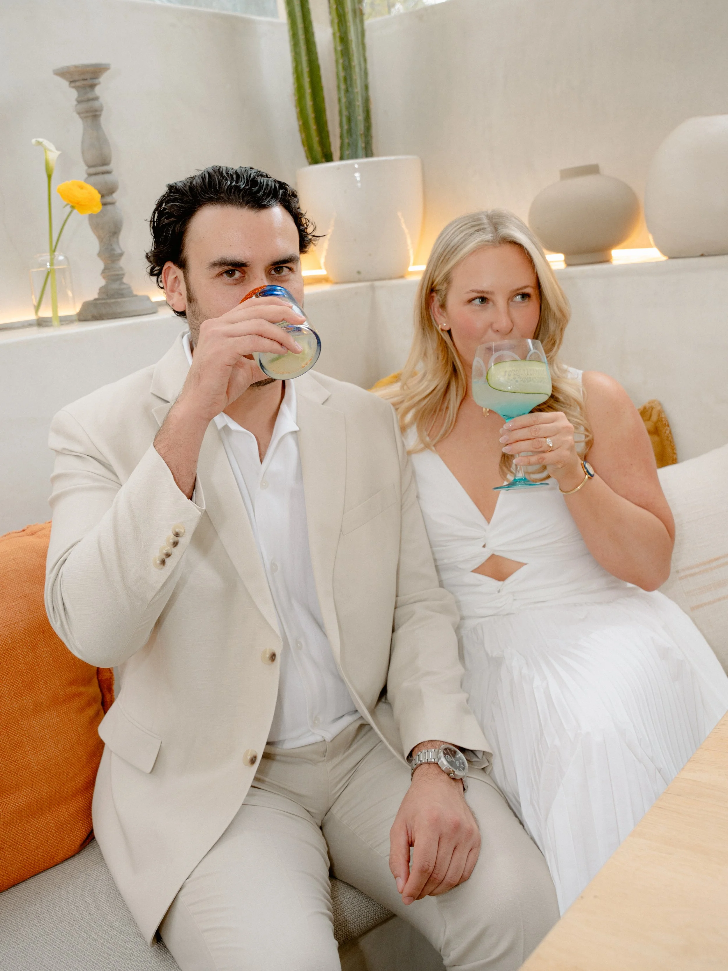 A man and woman sitting on a beige sofa, drinking cocktails with lime slices. The man has dark hair and is dressed in a cream suit, while the woman has blonde hair and is wearing a white dress. They are in a well-decorated room with plants and vases 
