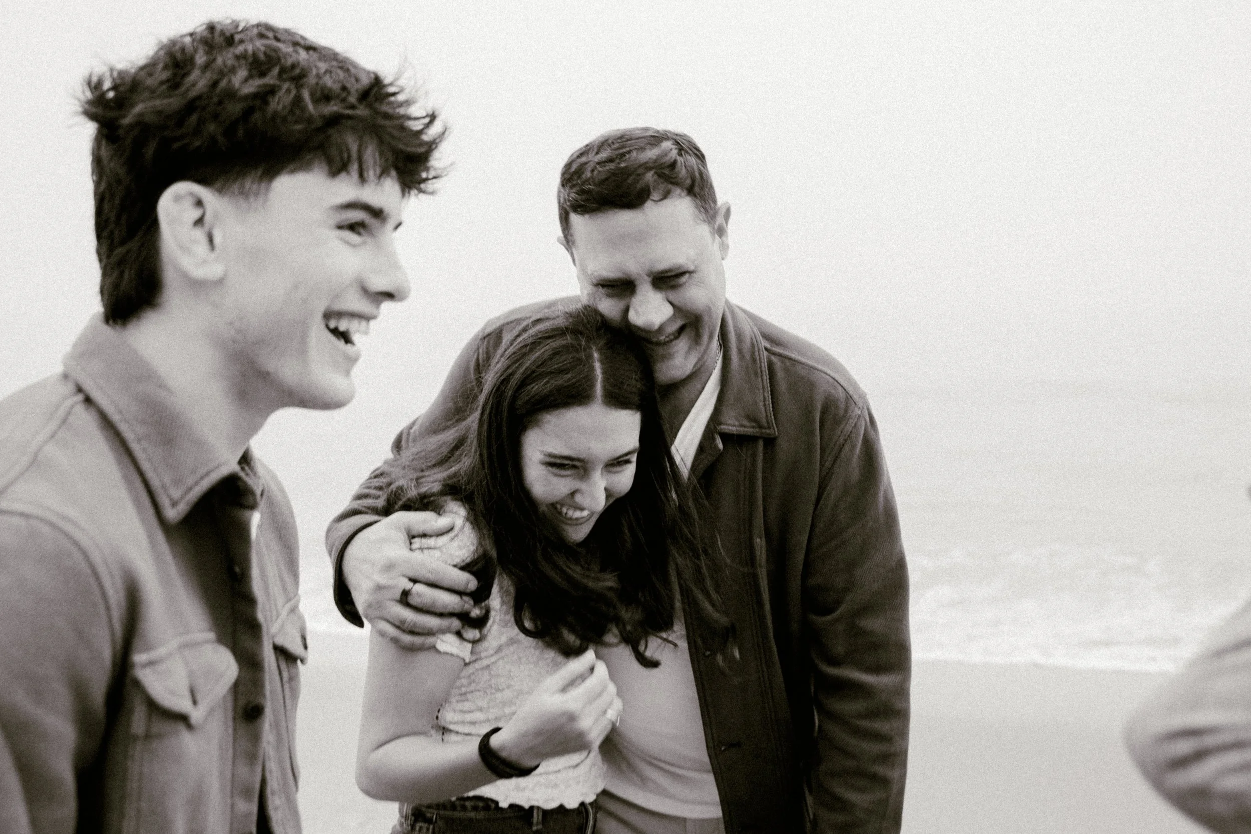 Four young people standing closely together outdoors, smiling and hugging, seen in black and white.