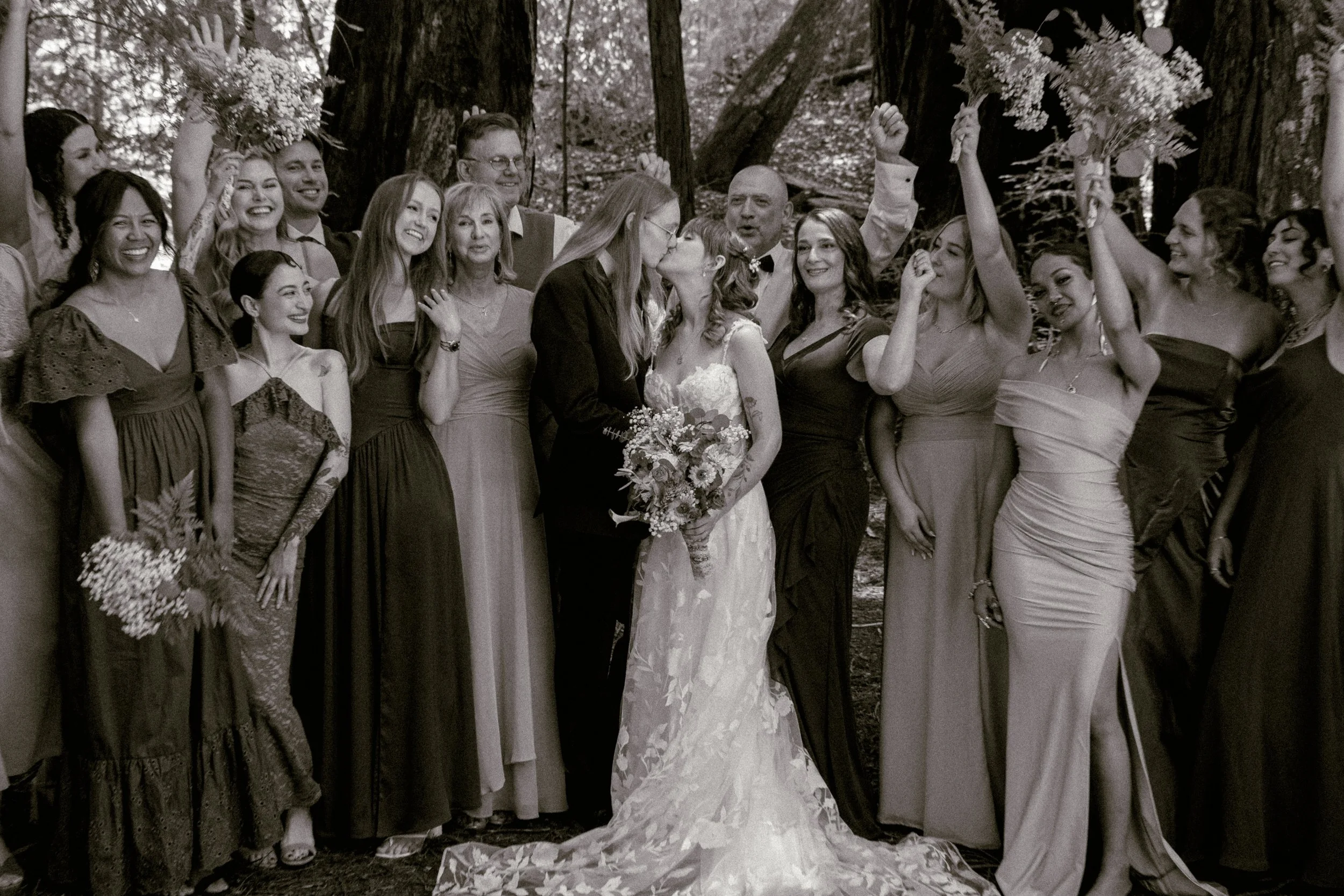 A wedding scene in a forest with the bride and groom kissing surrounded by guests who are smiling and celebrating.