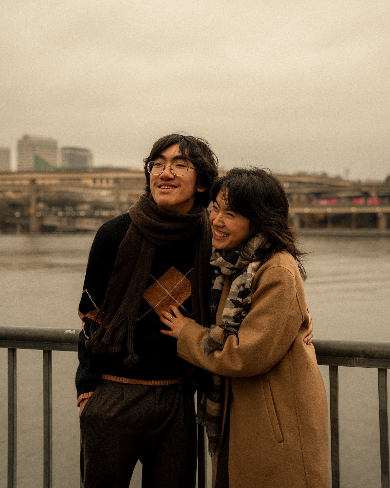 A man and a woman are standing close together by a river with a city skyline in the background. They are smiling and appear happy.