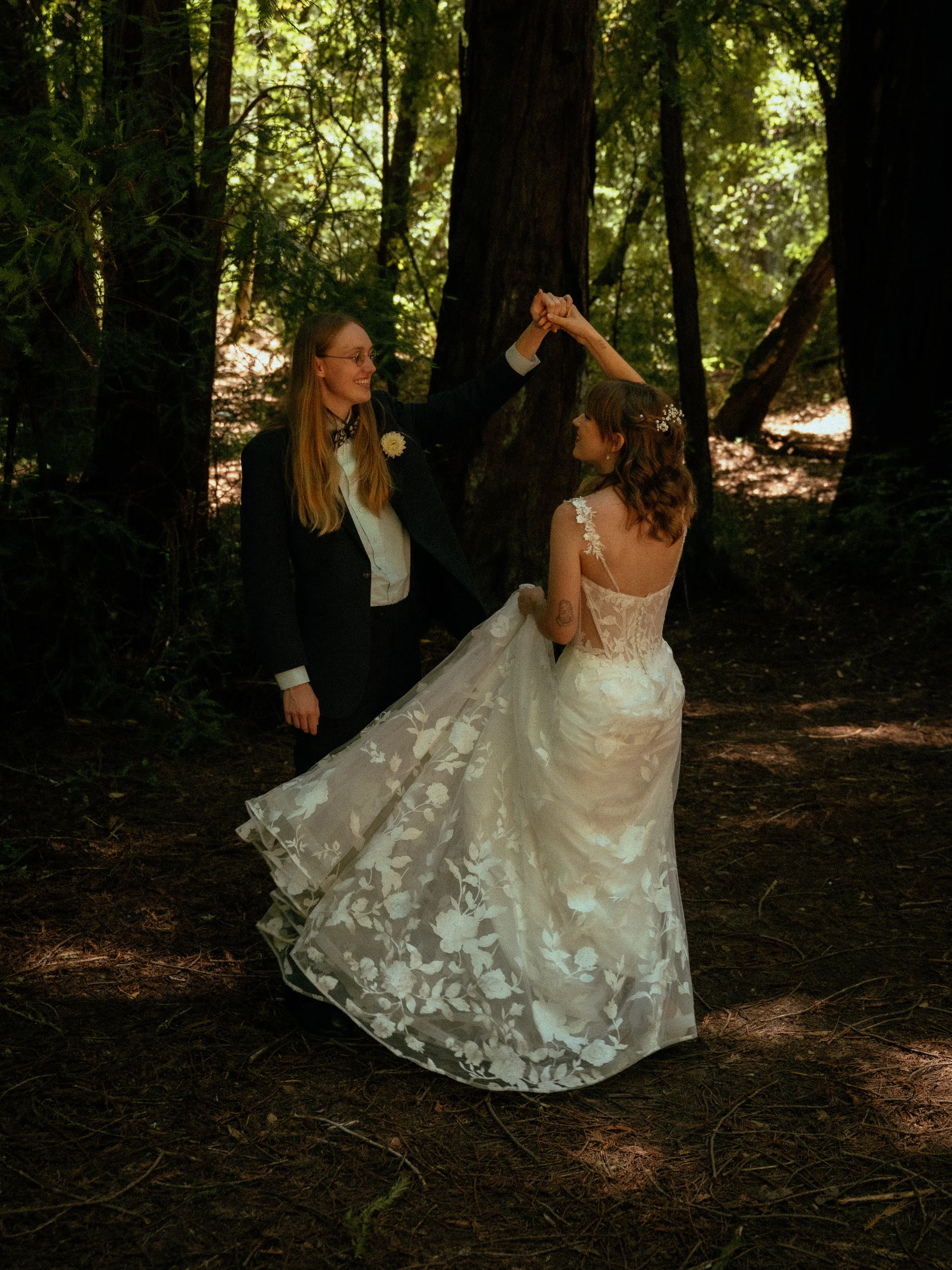 A couple dressed in wedding attire holding hands and dancing in a forest with tall trees and sunlight filtering through the foliage.