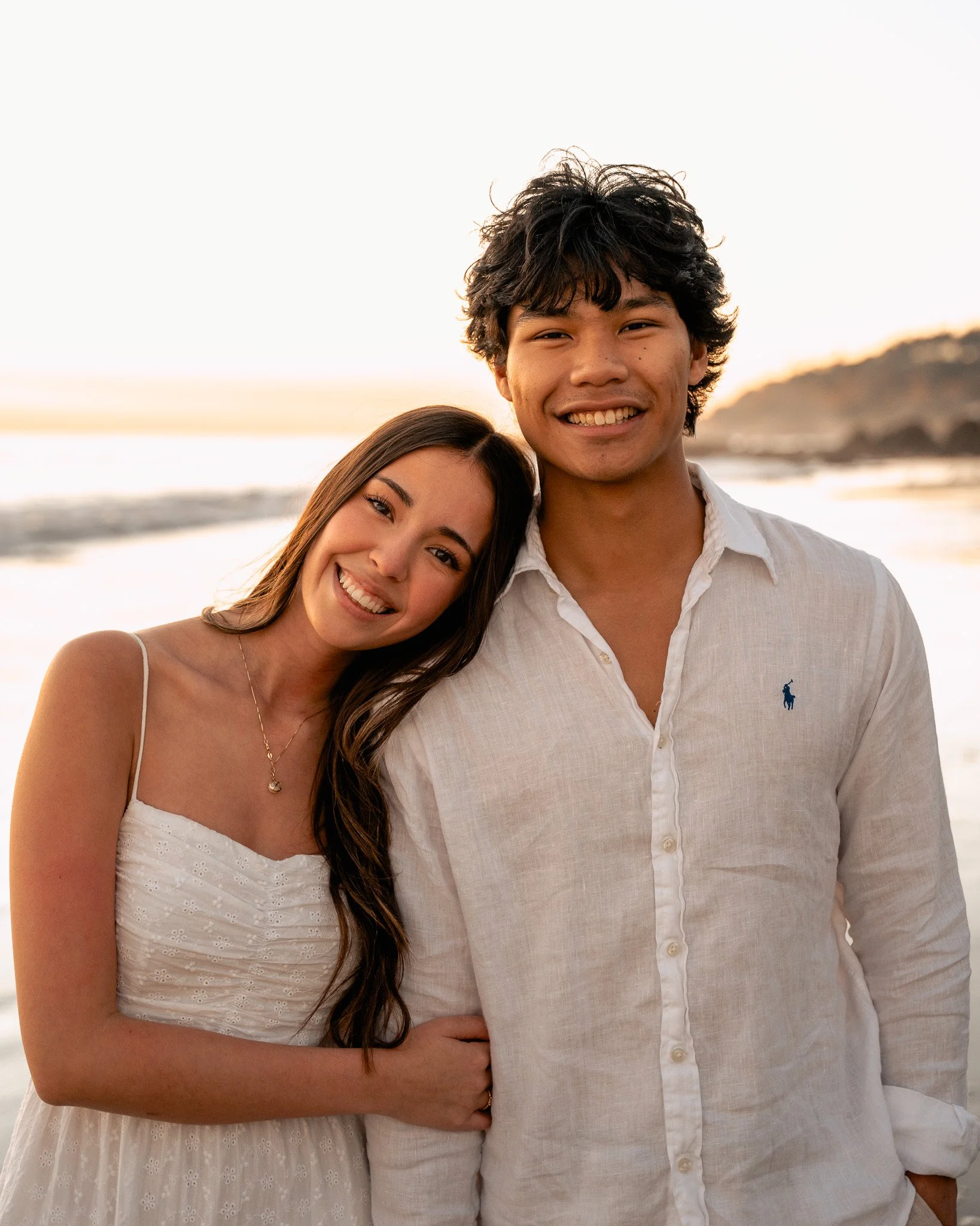 Close-up of a young couple smiling, standing near the ocean at sunset, with a blurry coastline in the background.