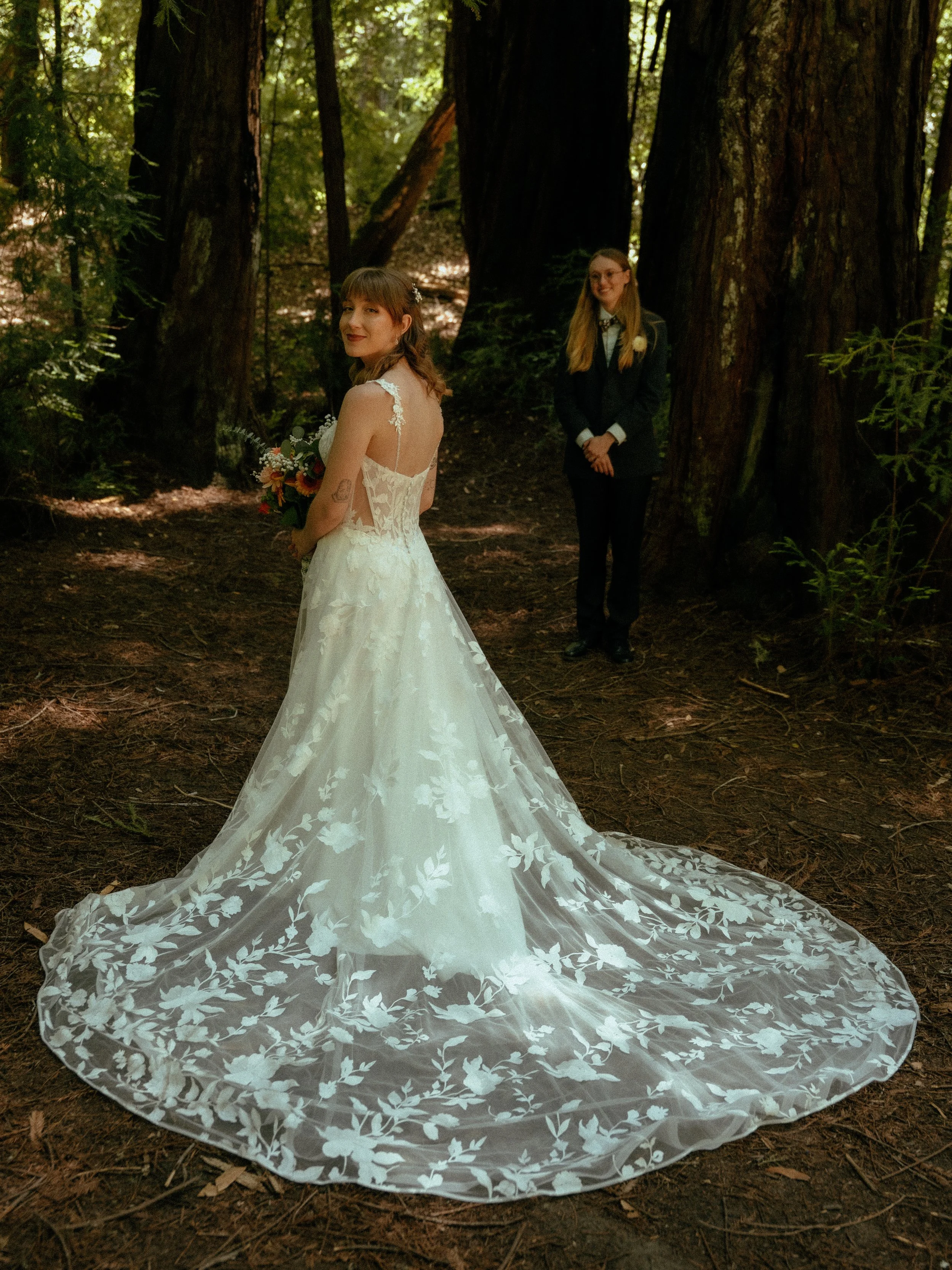 A bride in a lace wedding dress holding a bouquet, standing in a forest, with a bridesmaid in a tuxedo standing behind her and to the right.