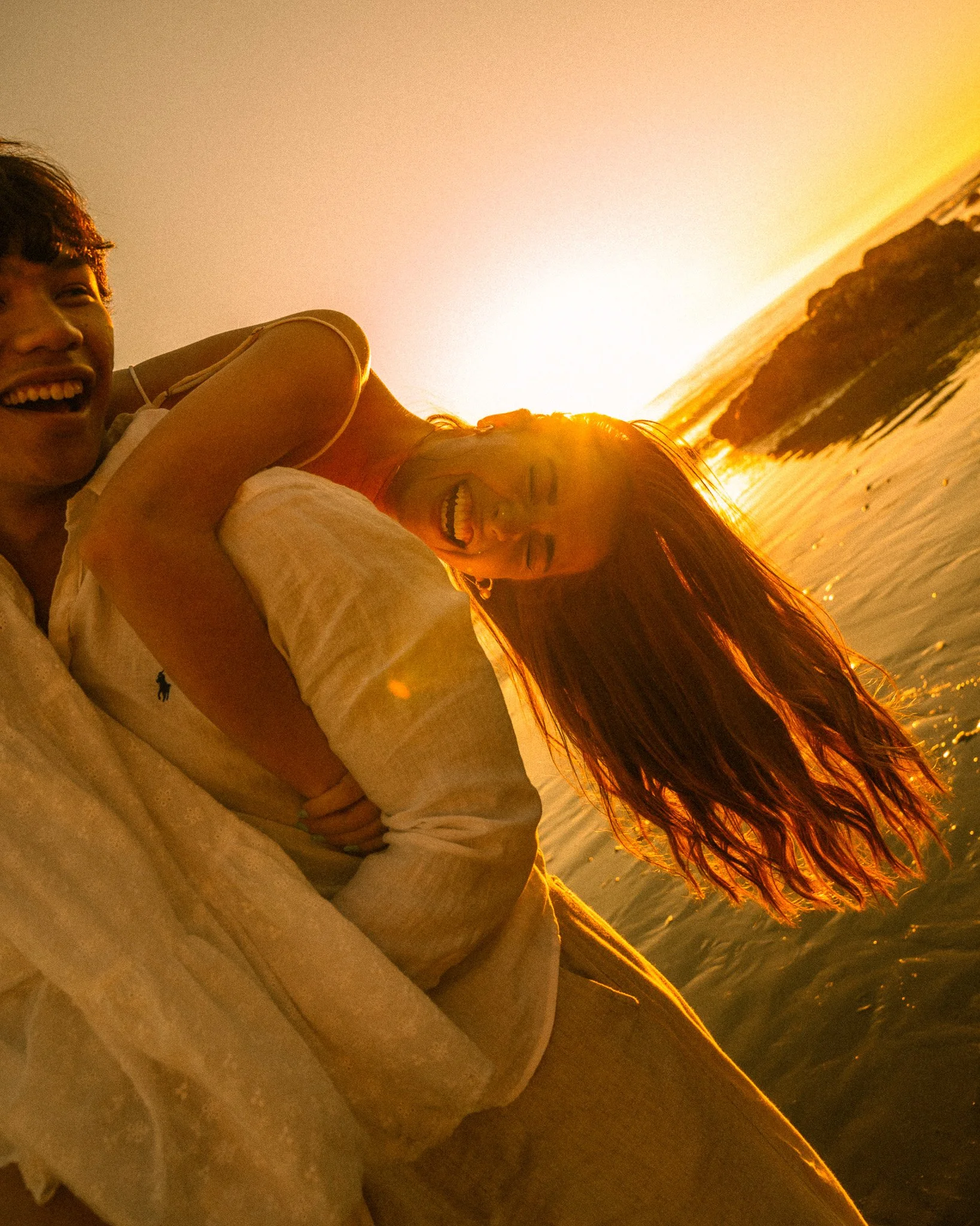 Two women smiling at the beach during sunset, with one wearing a beige shirt and the other with long, flowing hair.