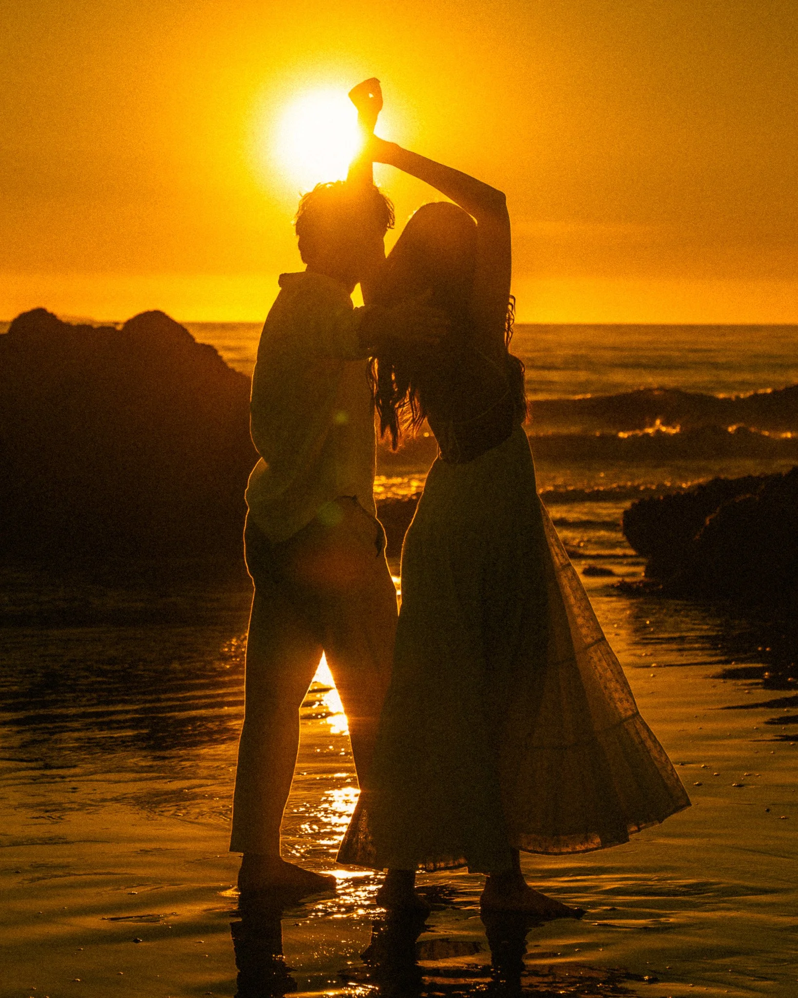 A silhouette of a couple standing close together by the ocean at sunset, with the sun setting behind them.