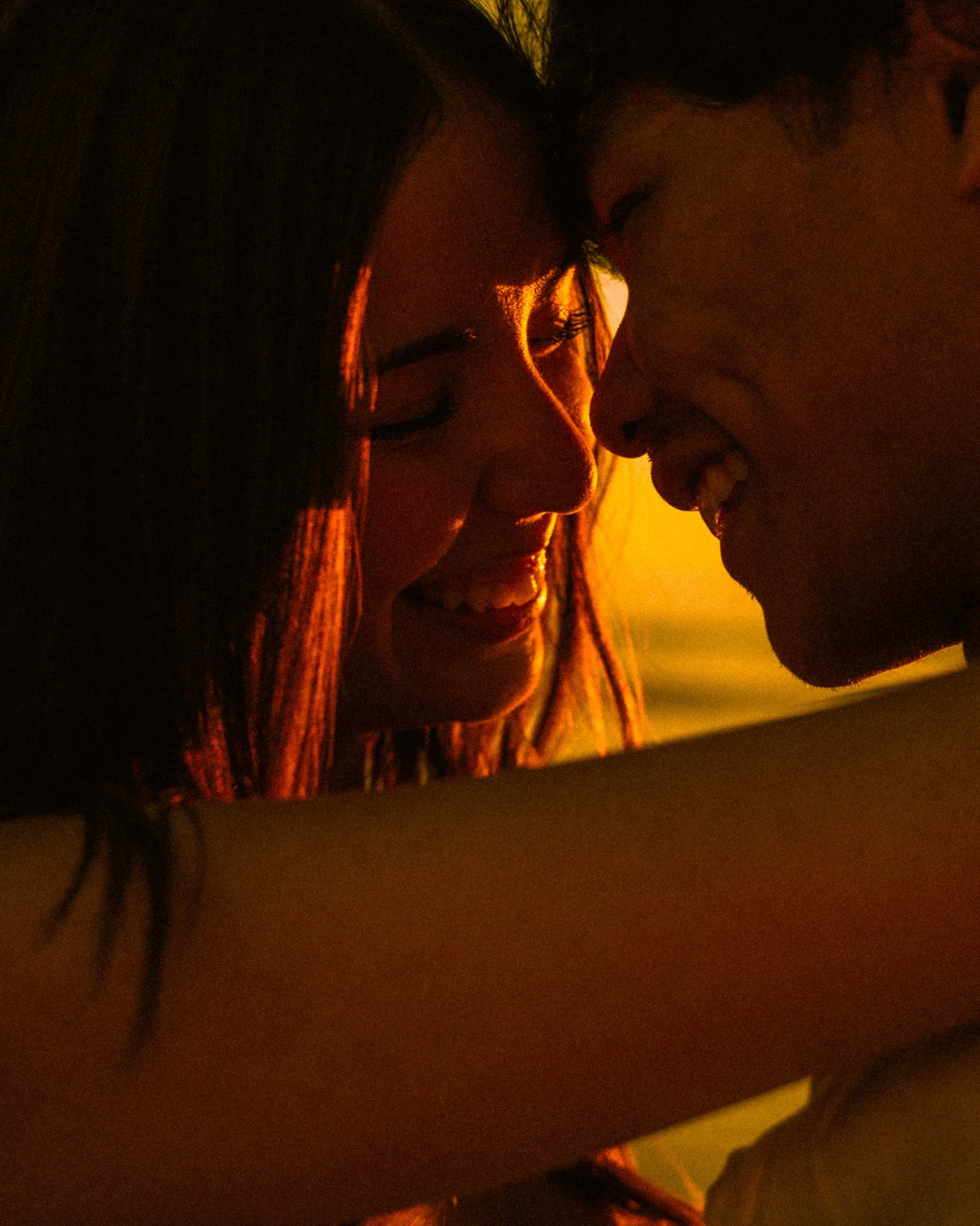 Two people are close together, with foreheads touching and smiling, during sunset or sunrise.