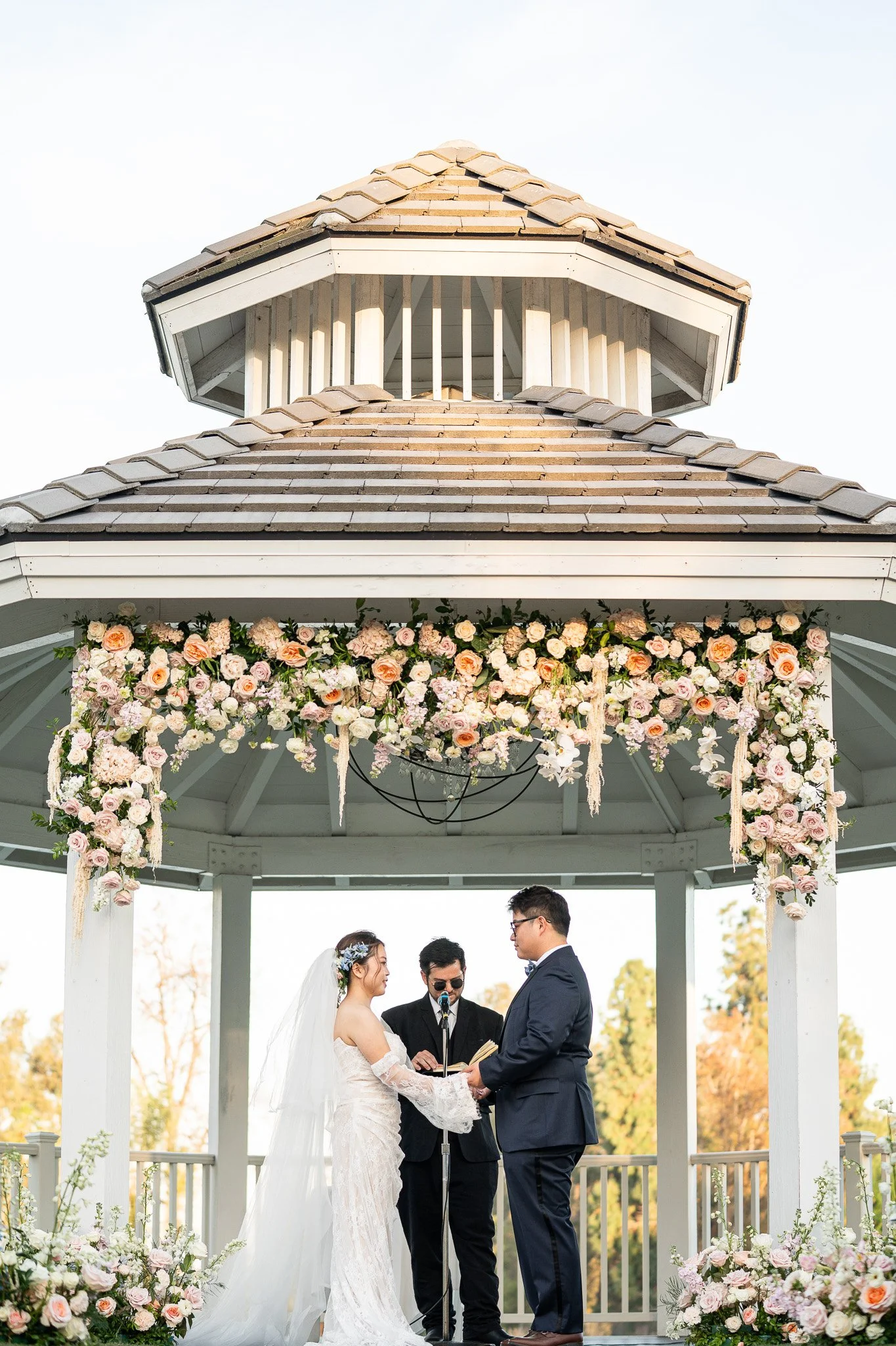A couple getting married outdoors in a gazebo decorated with pink and white flowers, with an officiant present.