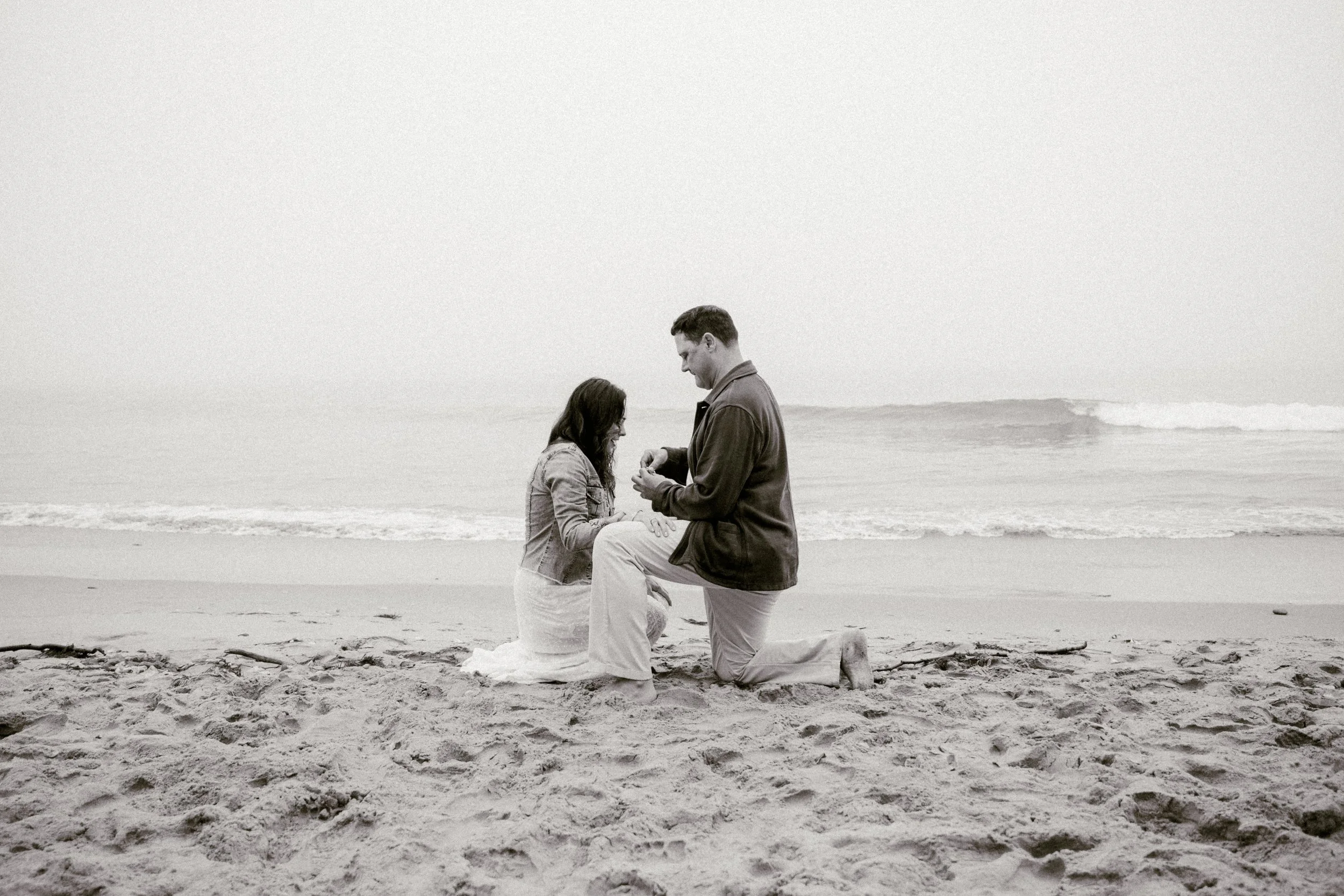 A black-and-white photo of a couple kneeling on the beach, with the man proposing to the woman. The woman is wearing a denim jacket and has long hair, while the man has short hair and is dressed in a jacket and pants. The ocean and waves are visible 