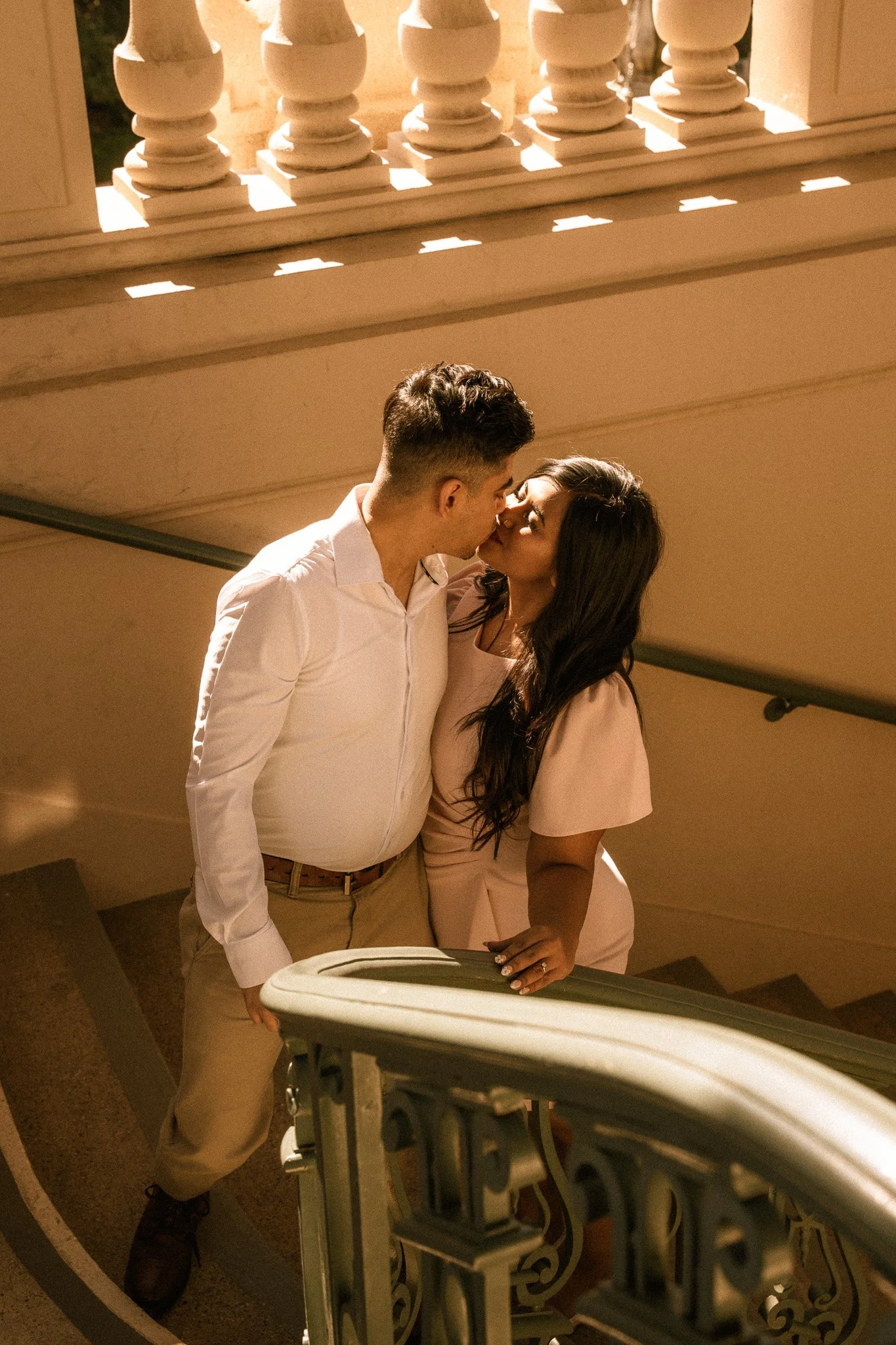 A couple kissing on a staircase under warm indoor lighting, with a white railing and a decorative arch in the foreground.
