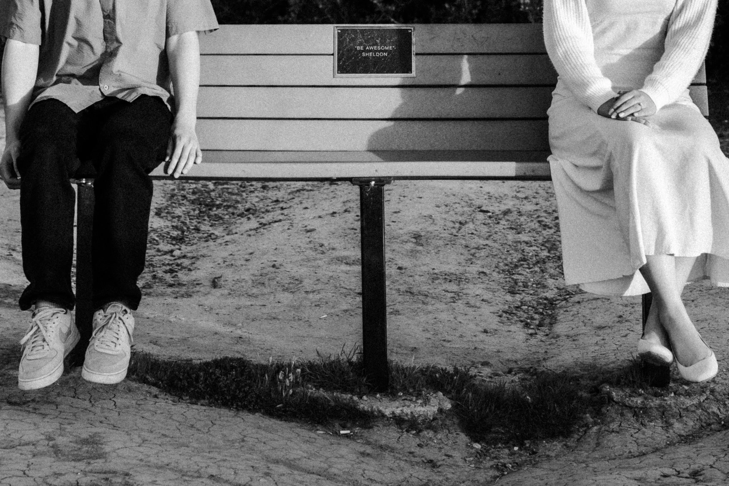 Black and white photo of a man and woman sitting on a park bench with a plaque that reads 'Be Awesome' Sheldon.