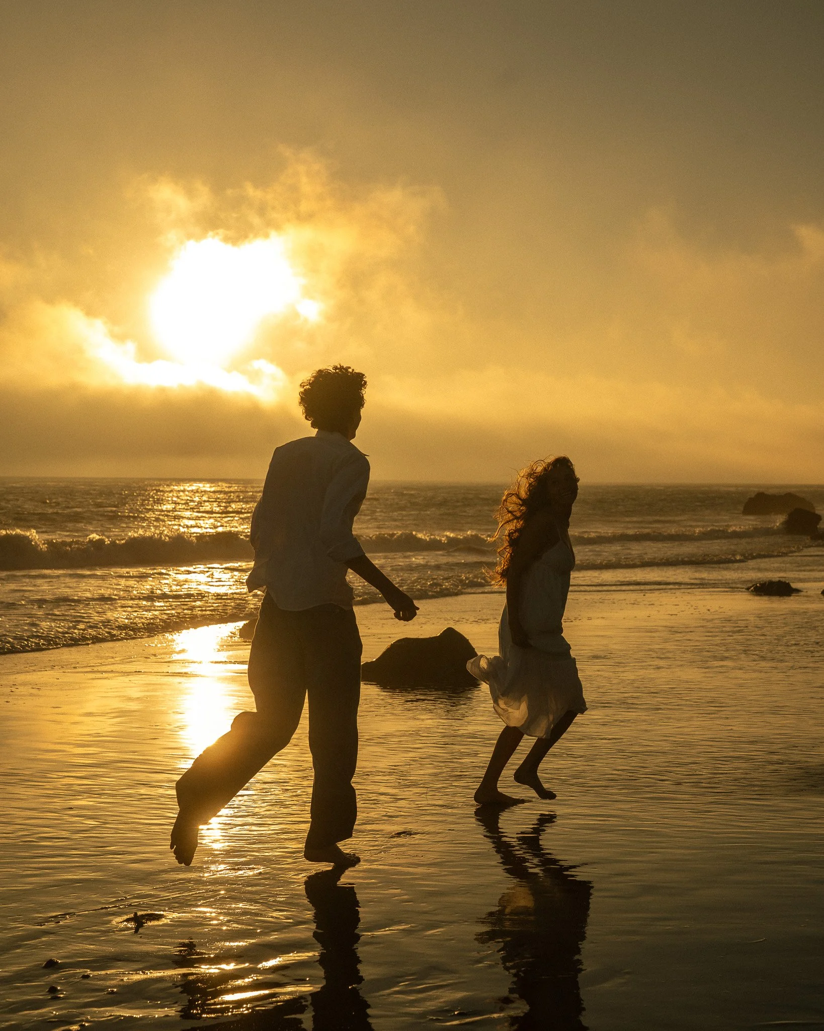 People are walking along the beach at sunset, with their reflections visible in the water.