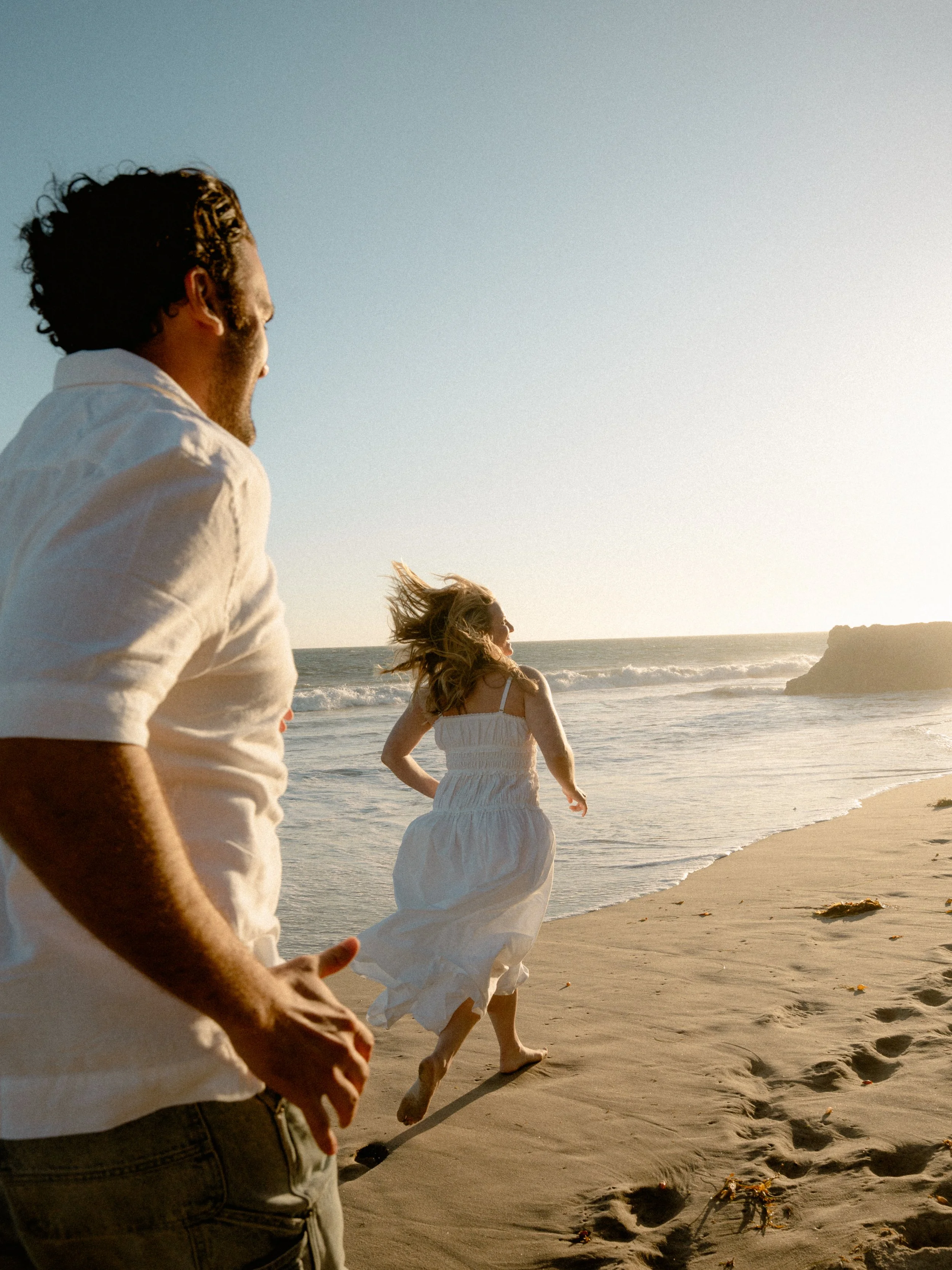 People walking along the beach during sunset or late afternoon, with the ocean and rocky formations in the background.