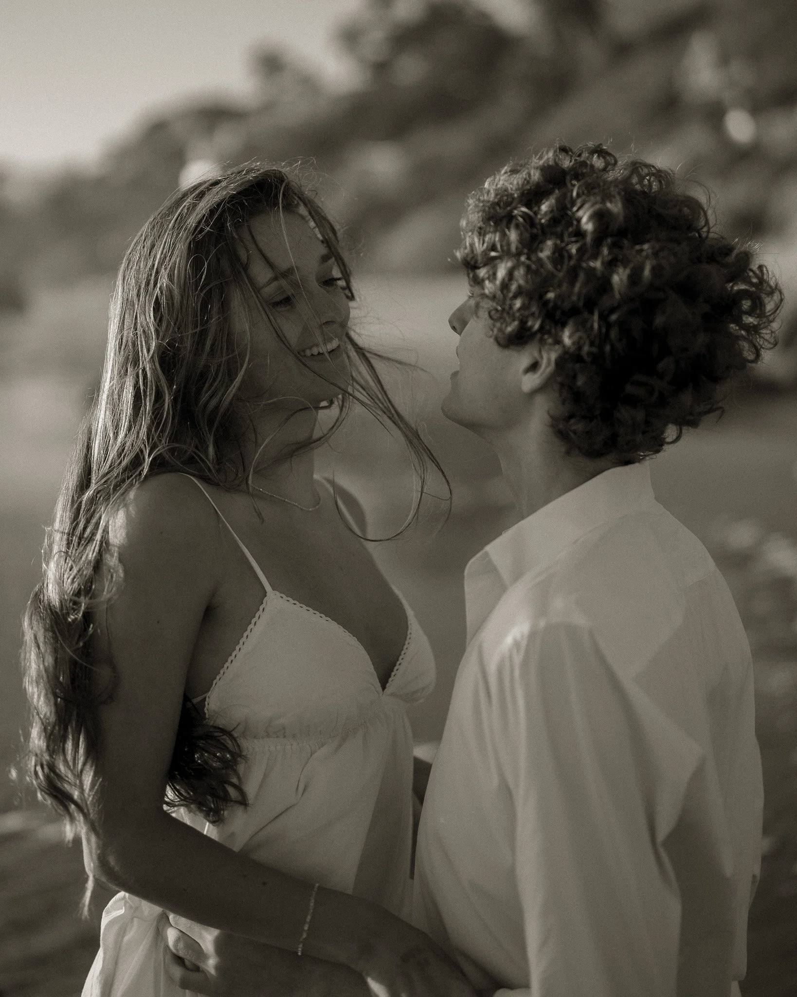 A black and white photo of a young woman and a young man on the beach, smiling and looking at each other closely, with the woman wearing a white dress and the man in a white shirt.