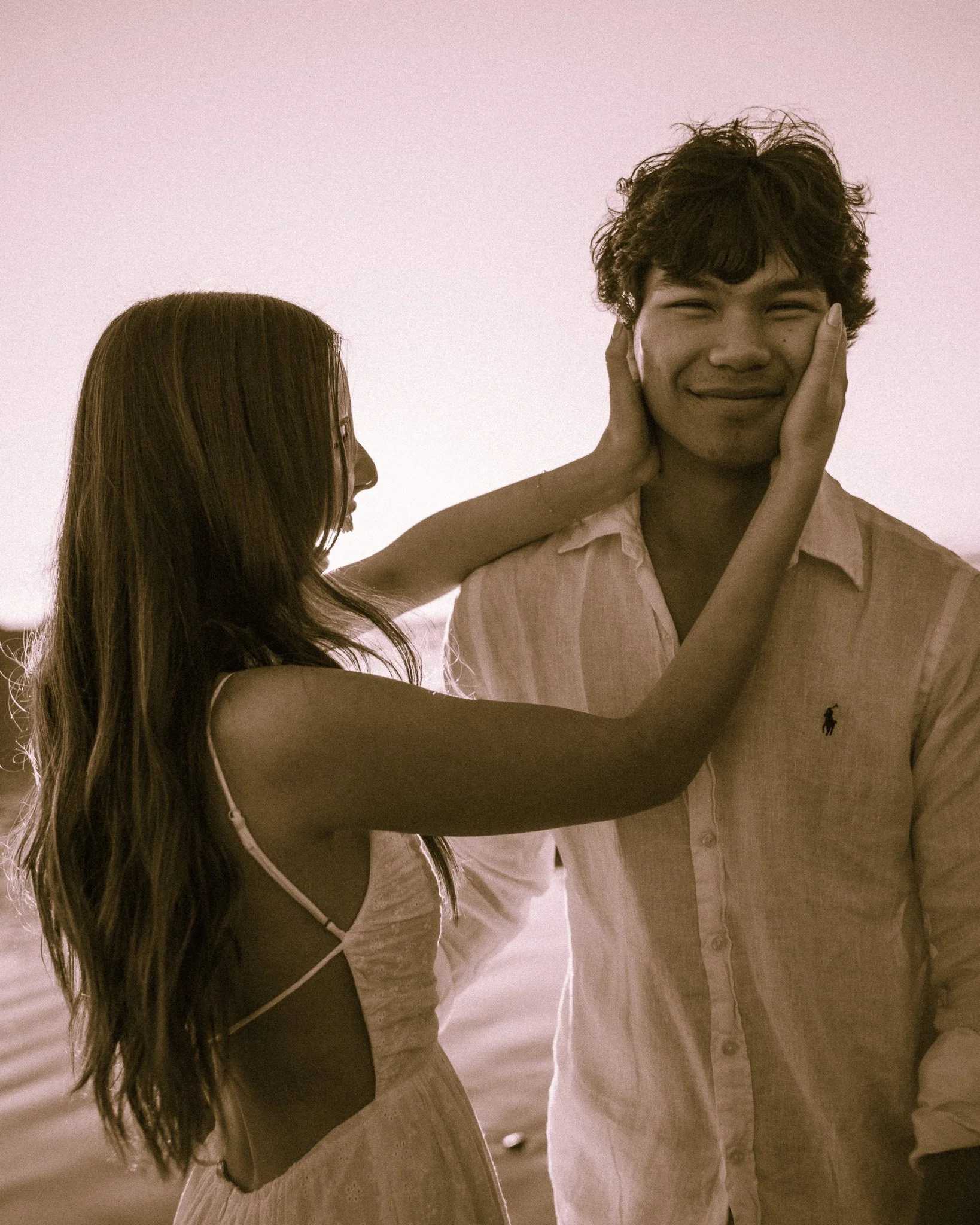 A woman playfully holds a man's face with both hands, and he smiles with his eyes closed. They are standing close to each other in a bright, softly lit room.