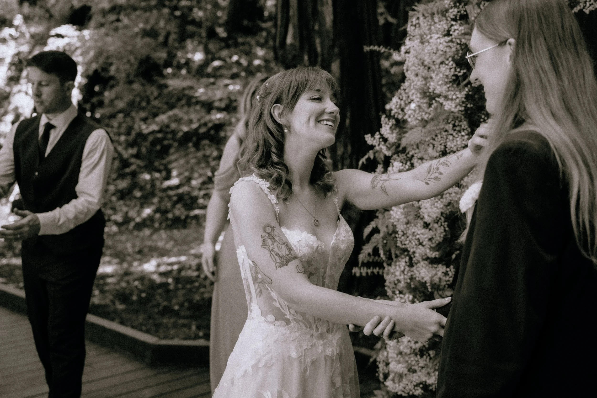 A bride and groom holding hands during a wedding ceremony outdoors, with a woman in a dress smiling at them, and a man in a vest and tie standing nearby.