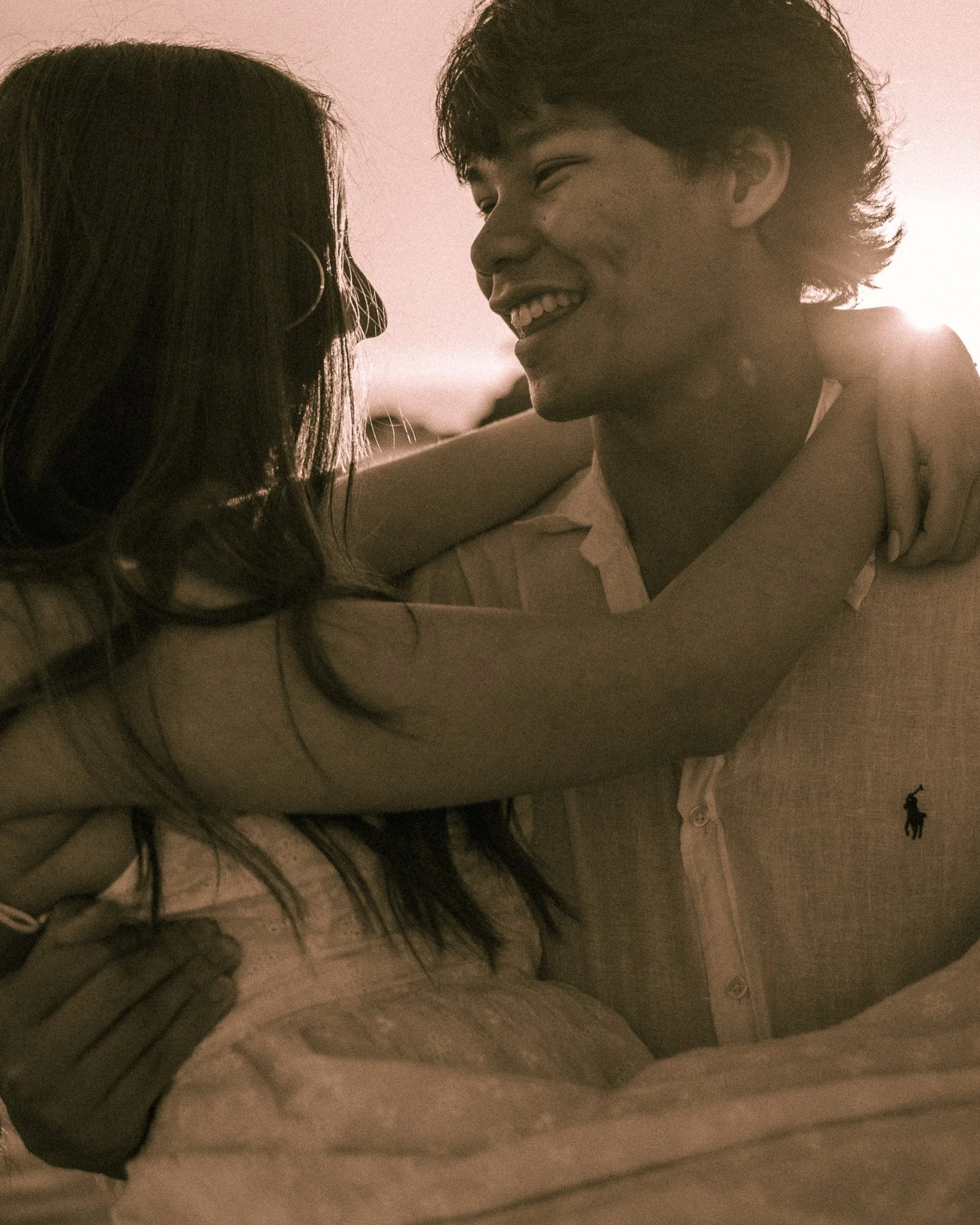 A young man and woman face each other closely, smiling and making eye contact in a warm, intimate moment. The woman has long dark hair and wears hoop earrings. The man has medium-length, curly hair and is dressed in a collared shirt. They are indoors