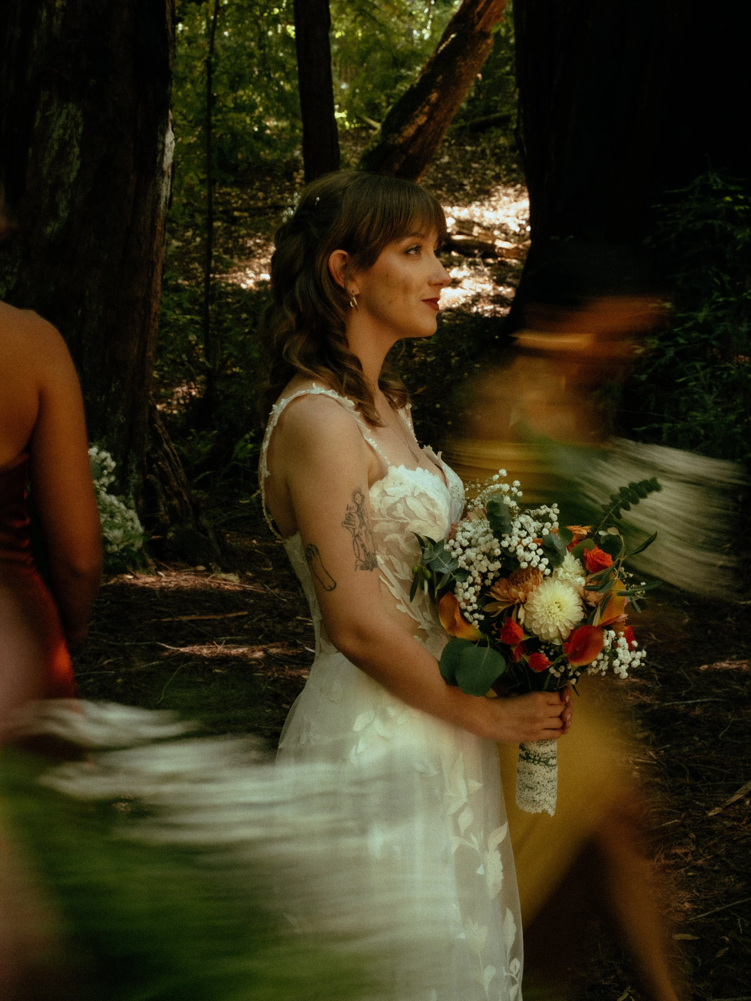 A woman in a white dress holding a bouquet of flowers stands in a forest. She has shoulder-length hair and tattoos on her arm, and the scene has a warm, natural lighting.