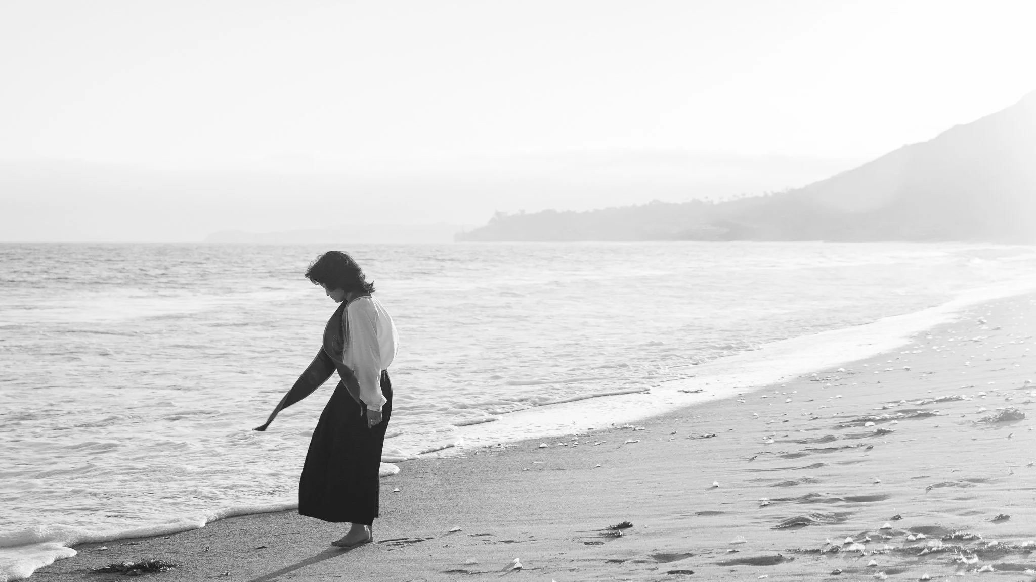 A woman in a traditional Japanese kimono walking barefoot along a beach with gentle waves lapping at her feet and a mountain in the background.