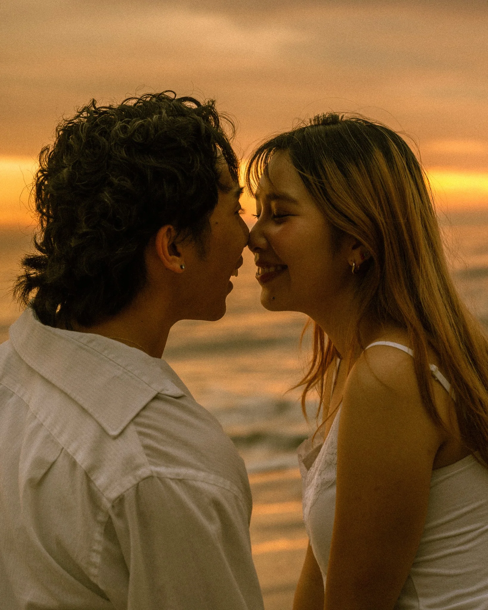 A close-up of a couple about to kiss at sunset, with the ocean and cloudy sky in the background.