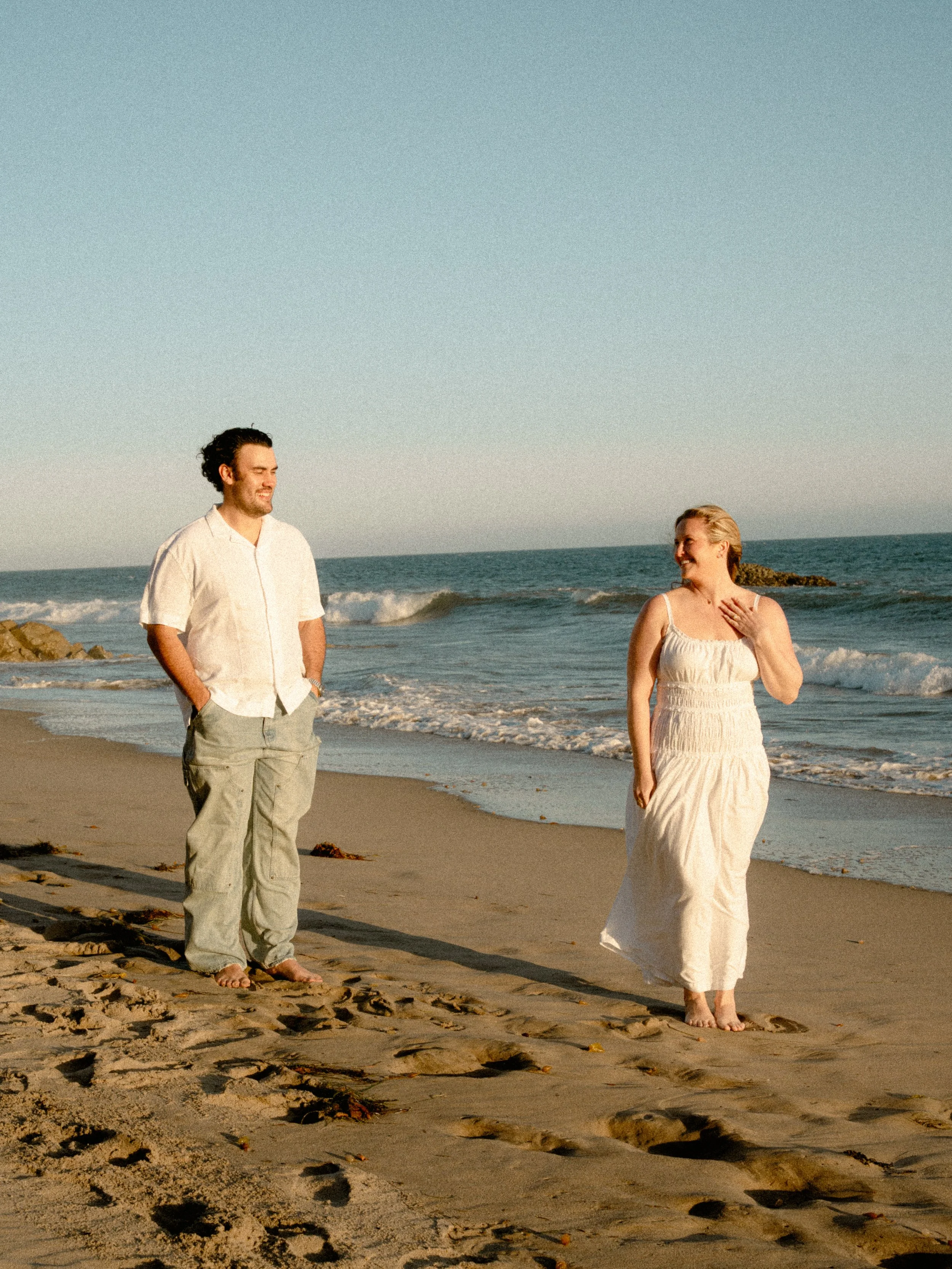 A man and woman walking on the beach by the ocean during sunset, with the man smiling and the woman touching her chest, both barefoot and dressed casually.