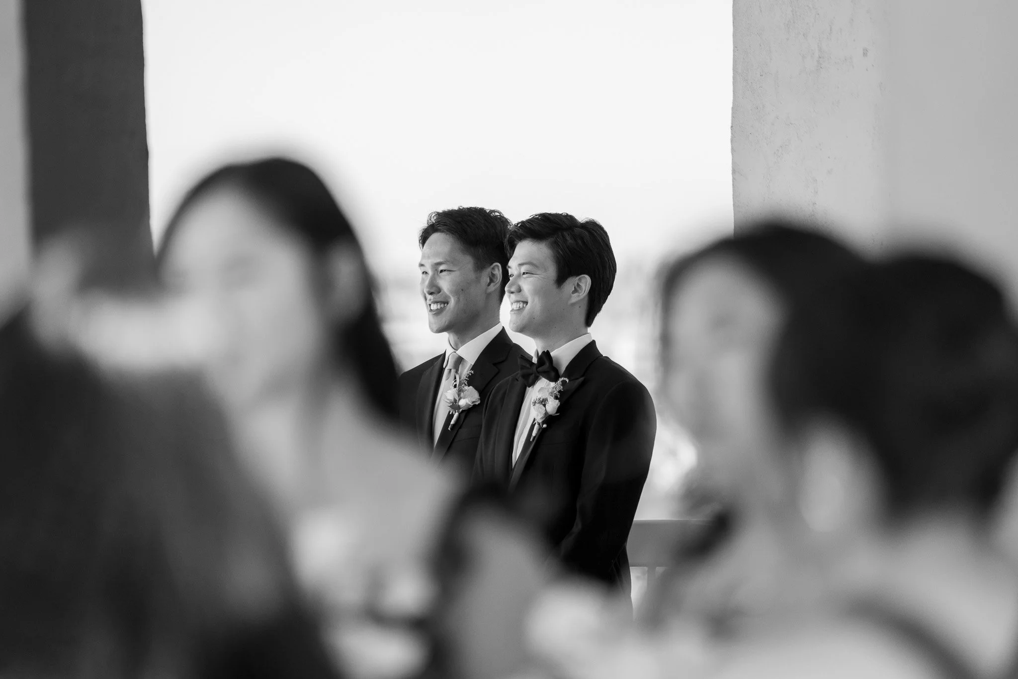 Black and white photograph of two men in tuxedos with bow ties, smiling and looking to the right, with blurred figures in the foreground.