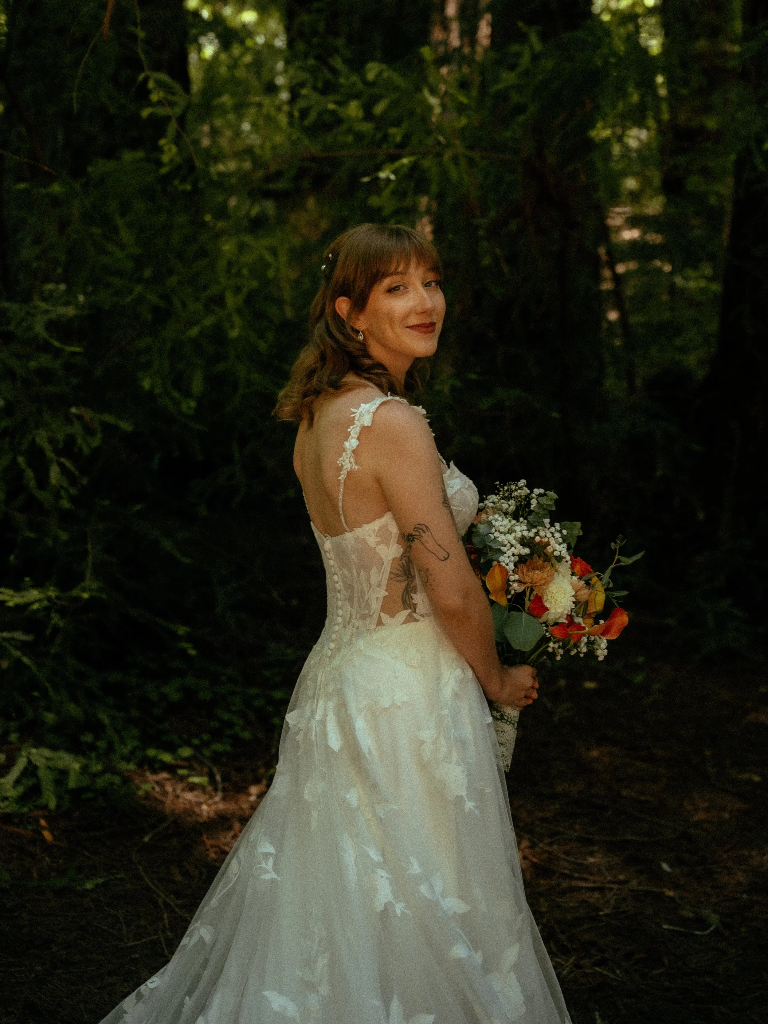 A woman in a white wedding dress holding a bouquet of flowers, standing in a forest with green foliage in the background, smiling at the camera.