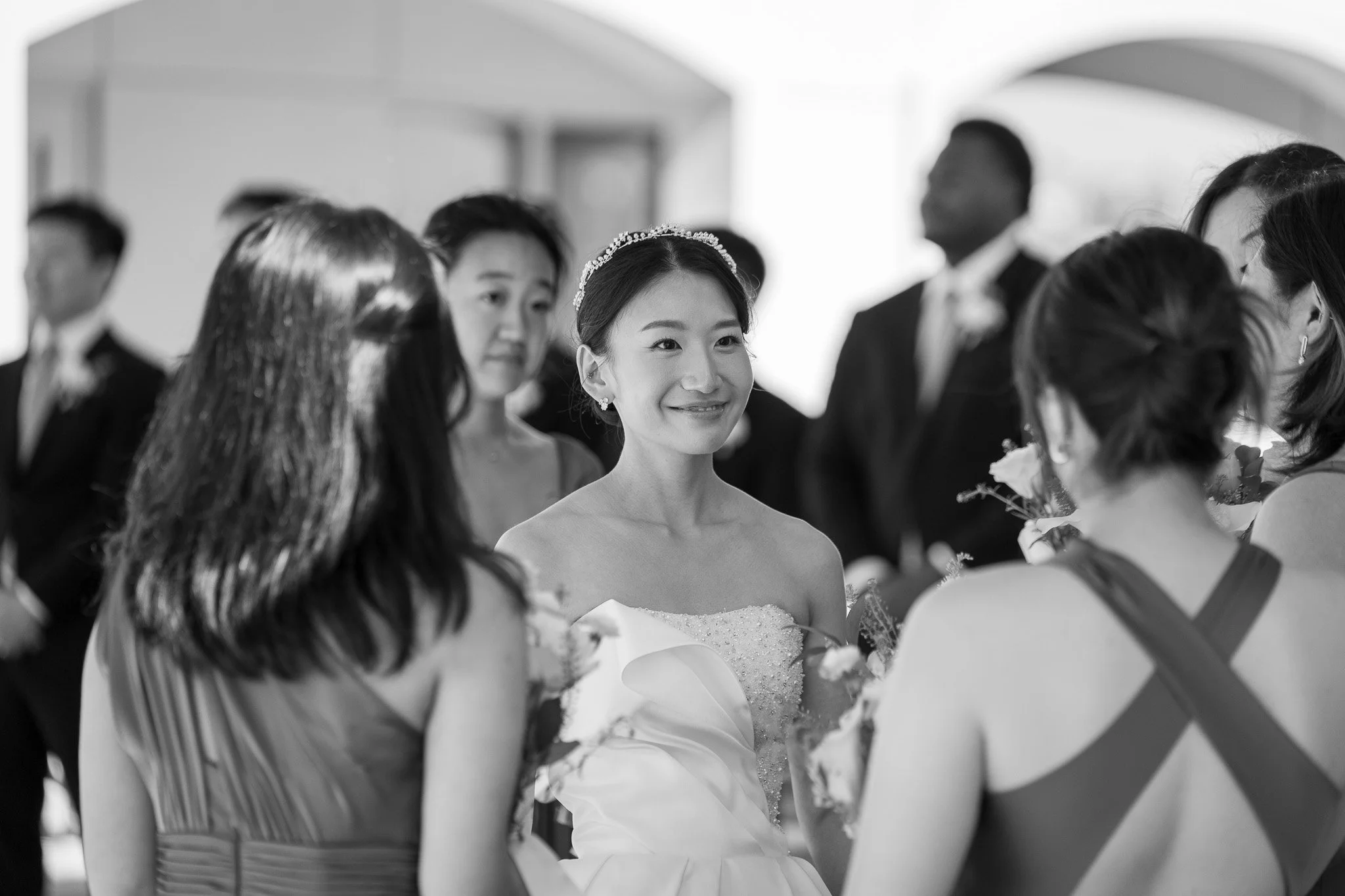 Black and white photo of a bride with a tiara on her head, smiling, surrounded by women and men at a celebration.