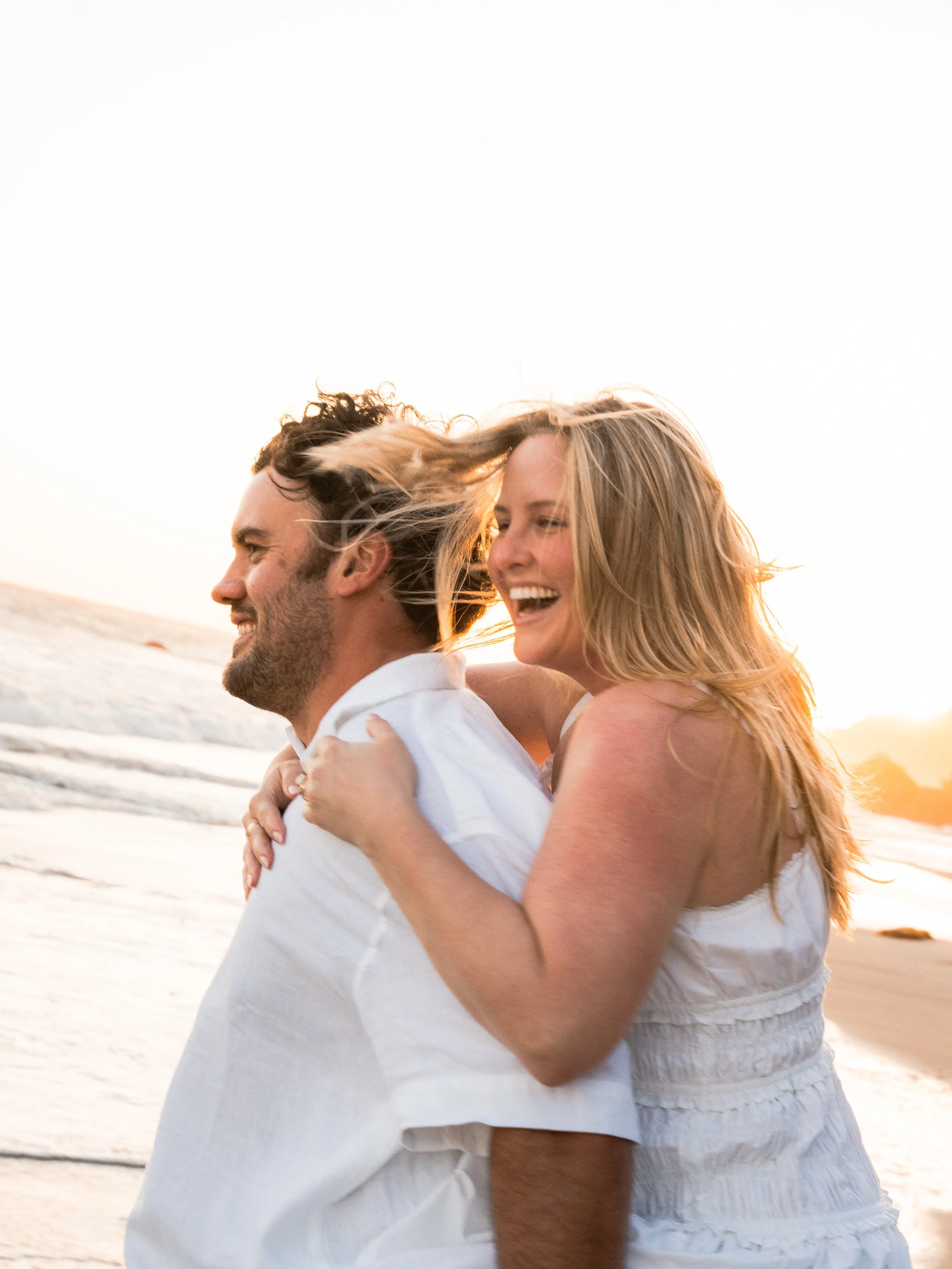 A smiling couple enjoying a walk on the beach at sunset, with the ocean and horizon in the background.